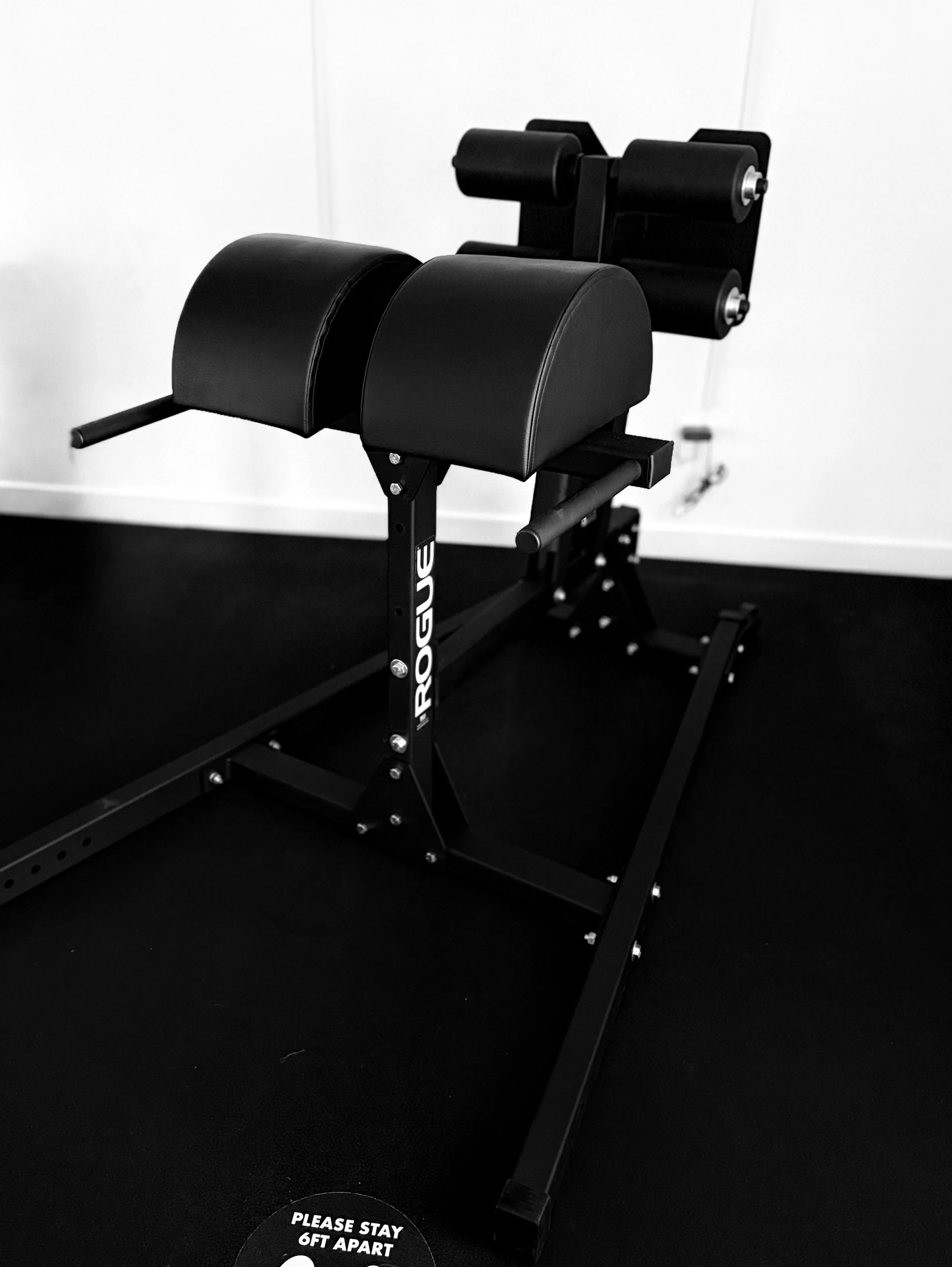 A black and white draft artistic style image of a leg curl strength training machine, with padded supports for the legs and adjustable components, set against a plain wall. There is a circular sign on the floor that reads, "Please stay 6 ft apart."