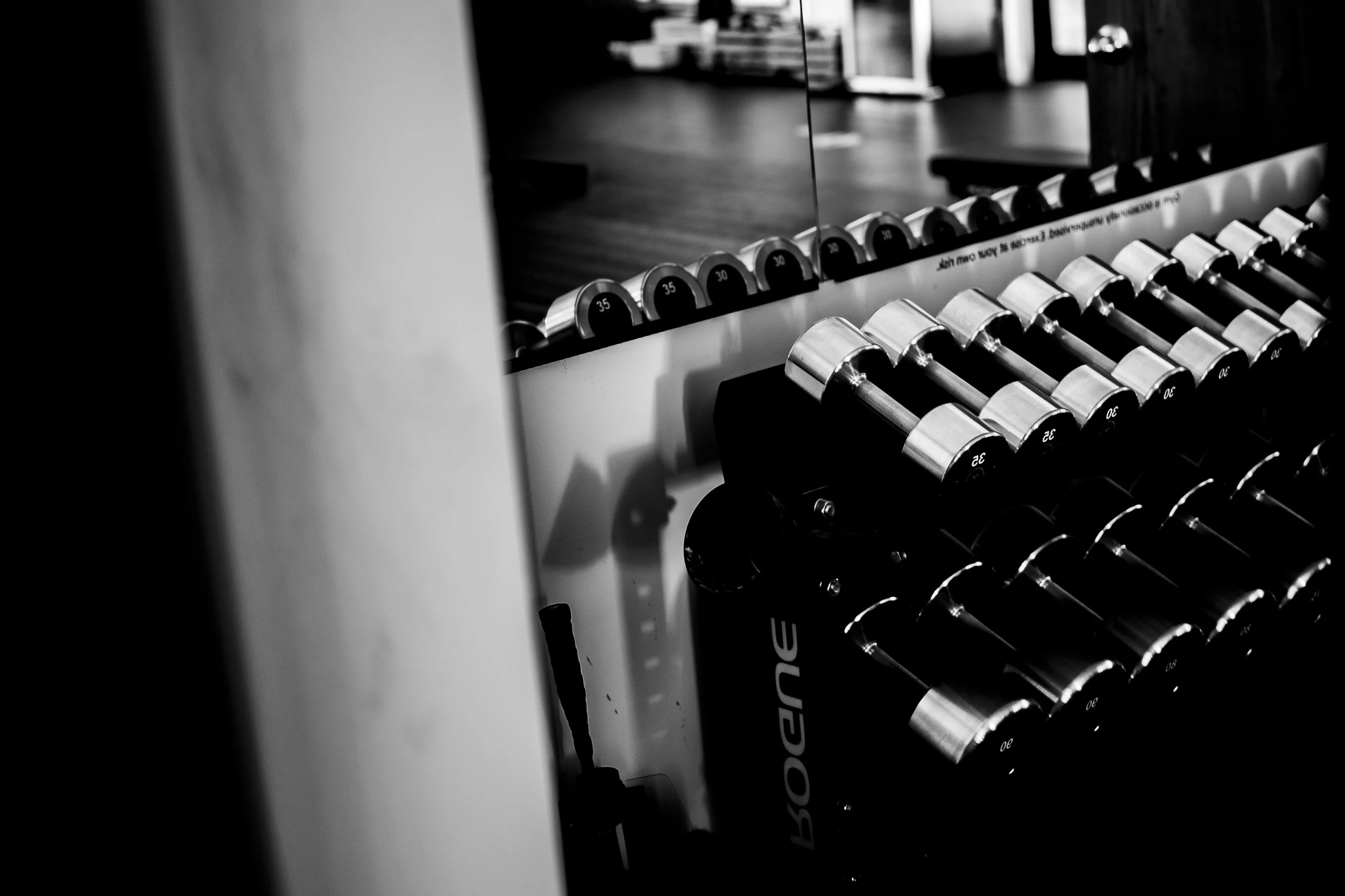 A black and white photo of a row of dumbbells on a rack, with a mirror reflecting part of a room in the background.