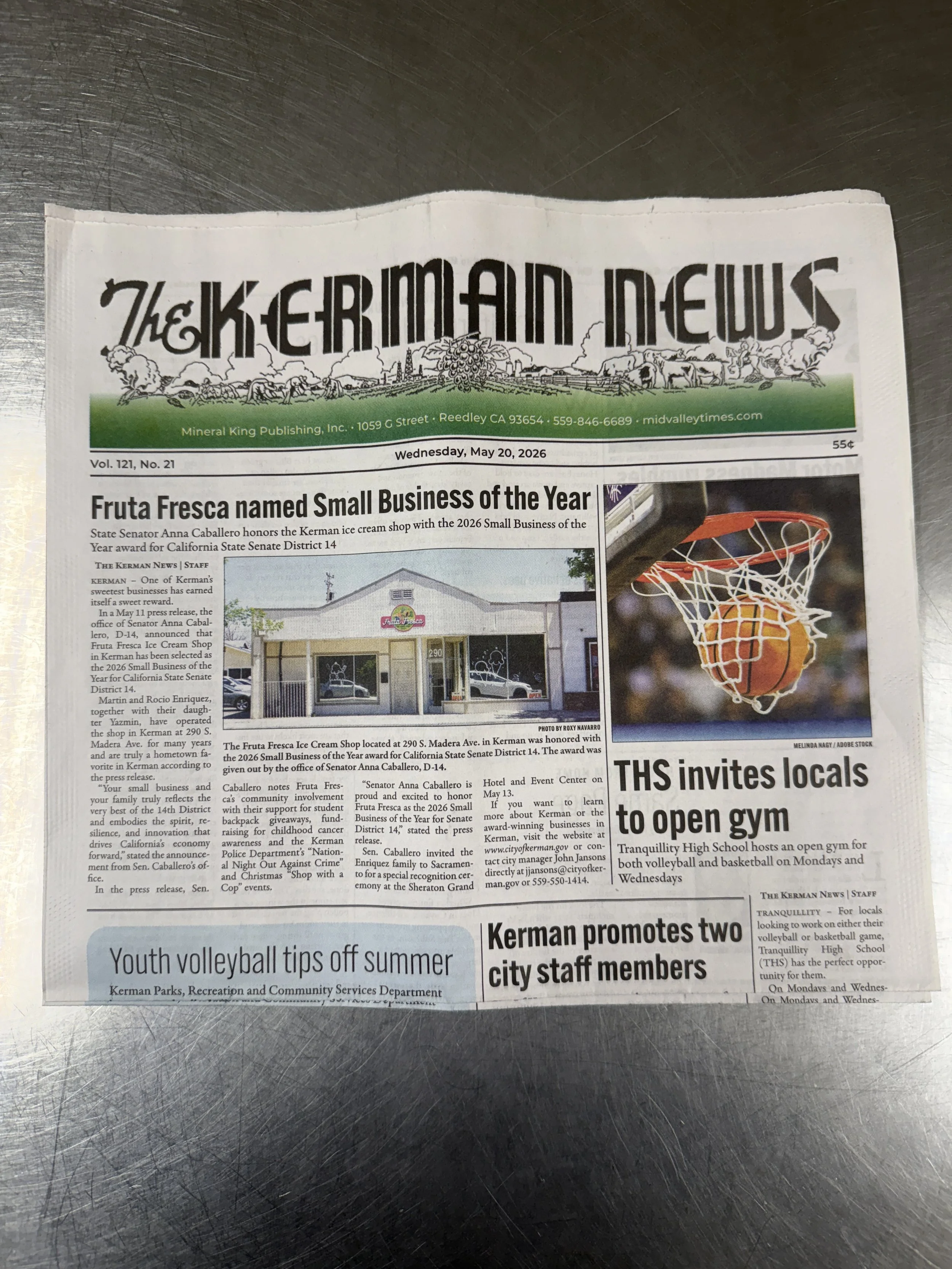 Front page of a newspaper titled "The Kerman News" with headlines about a small business award for Fruta Fresca, a photo of the shop, a basketball hoop with a ball going through it, and articles on local events and community updates.