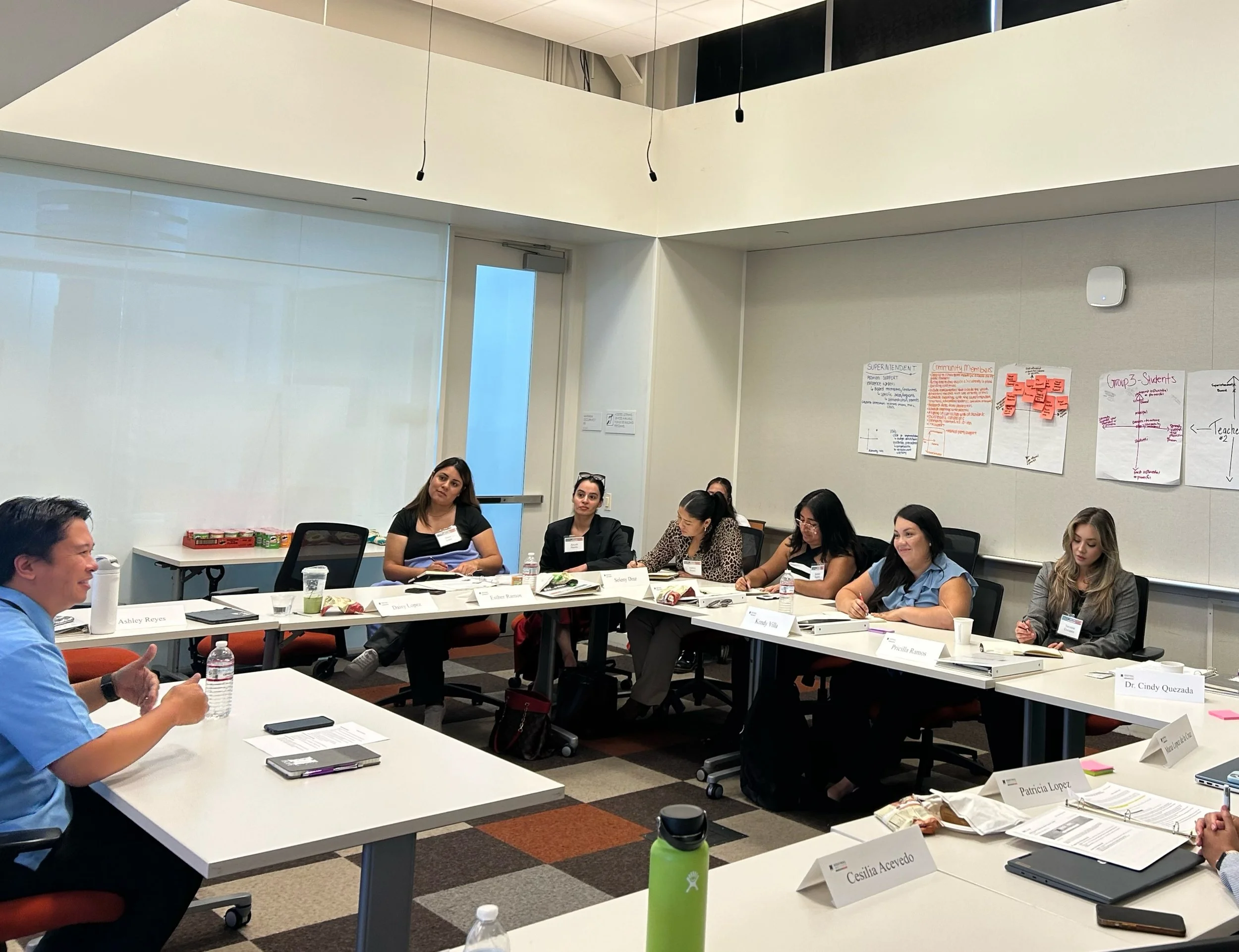People attending a business meeting or workshop in a conference room, seated around tables with name tags, notebooks, and water bottles, with presentation posters on the wall.