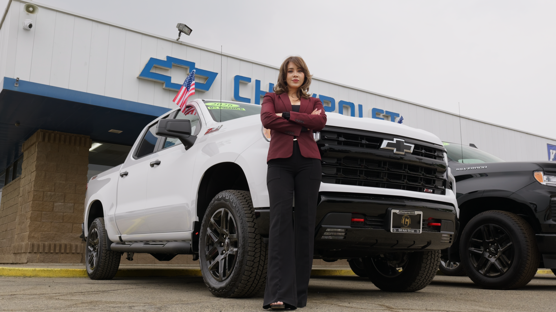 in a burgundy blazer and black pants standing with crossed arms in front of a white Chevrolet Silverado pickup truck outside the Gill Chevrolet dealership.