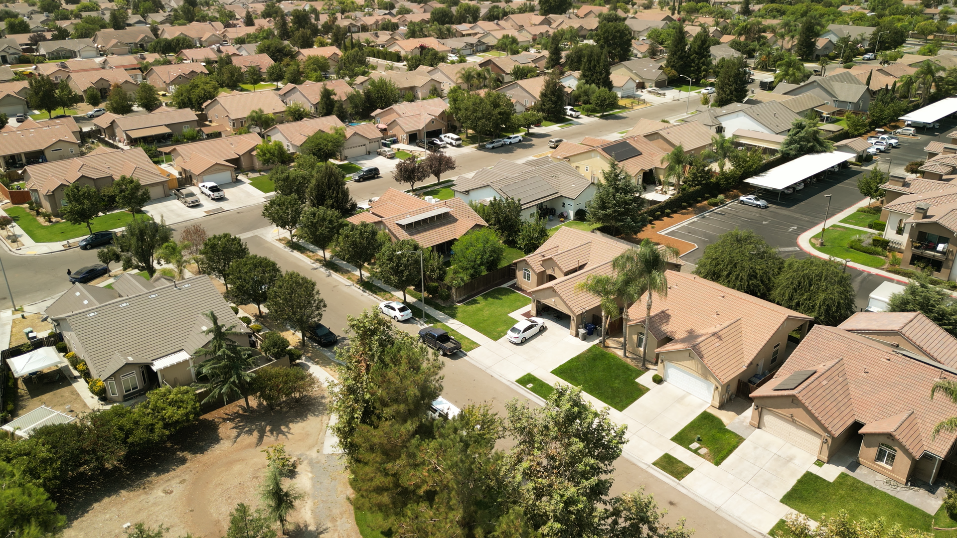 An aerial view of a Kerman, CA residential neighborhood with single-family homes, many with tiled roofs, surrounded by trees and greenery, with a few parked cars and a covered parking area.