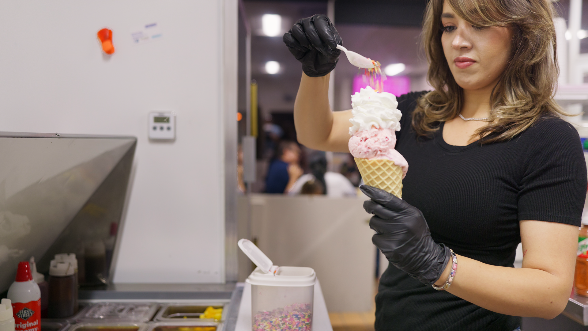 Yazmine Enriquez, 2026 Kerman Mayor Candidate decorating an ice cream cone with colorful sprinkles in a commercial kitchen or ice cream shop.