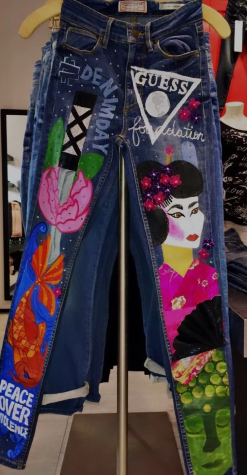 Pair of blue jeans with colorful painted designs, including a pink and green cactus, an orange and blue fish, a woman in traditional Japanese attire with flowers in her hair, the words 'GUESS' and 'DEN' and 'GAY' and other decorative elements.