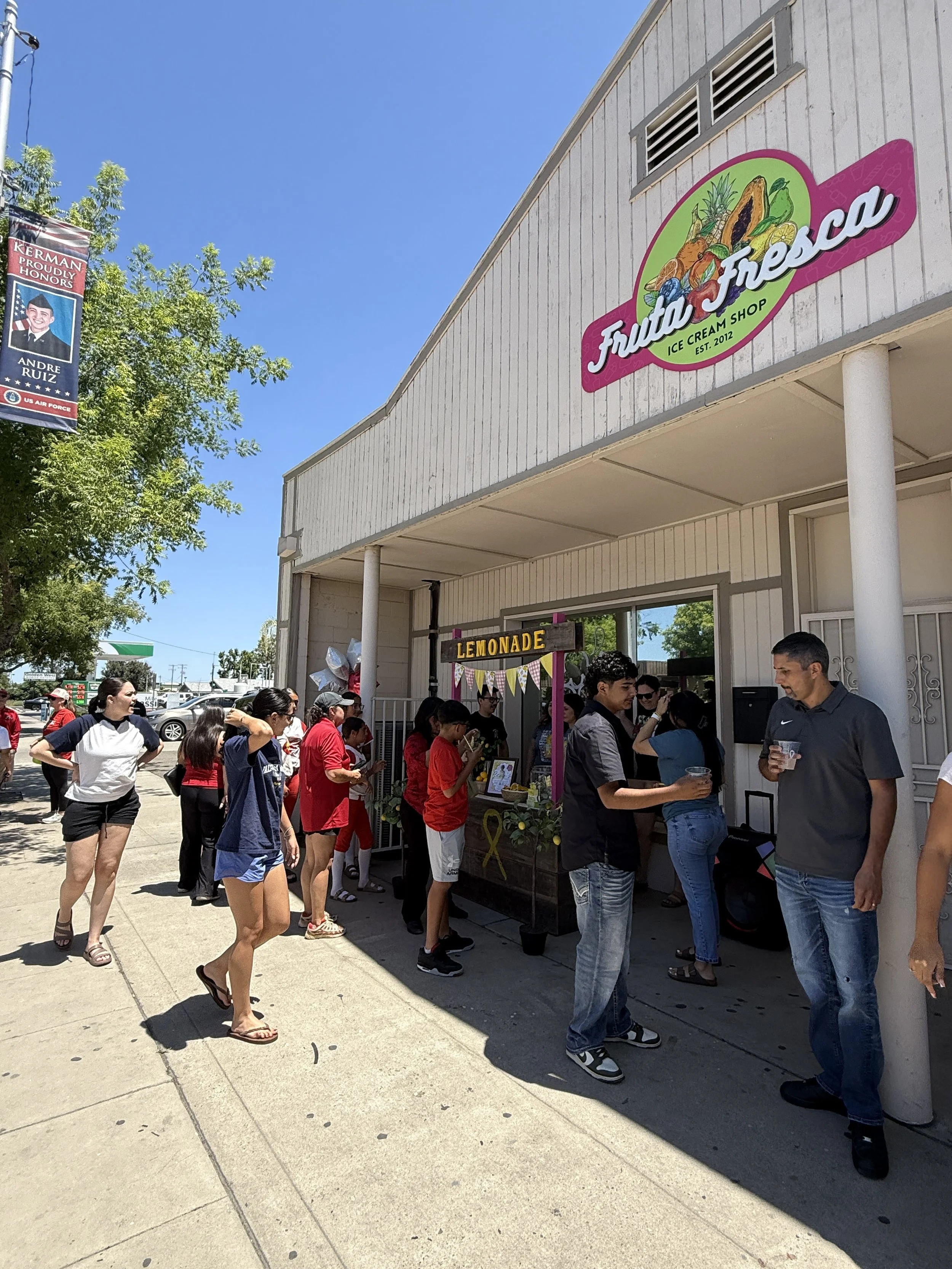 A group of people standing outside a storefront with a sign that reads 'Fruita Fresca Ice Cream Shop.' There is another sign below that says 'Lemonade.' The storefront has a porch with white columns, and there are trees and cars in the background on a sunny day.