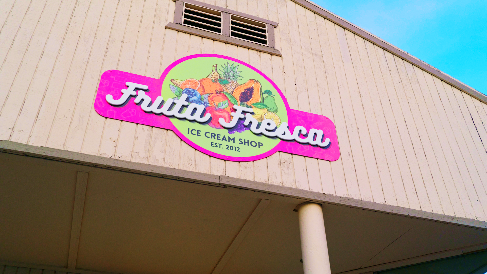 Sign for Fuda Fresa ice cream shop with colorful fruit illustrations and established in 2012, mounted on a beige wooden building with a vent near the roof and a blue sky above.