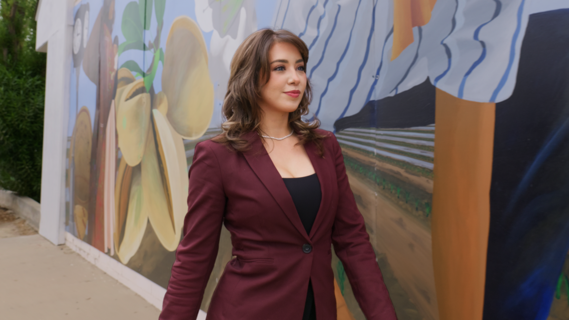 Yazmine Enriquez, 2026 Kerman Mayor Candidate in a burgundy blazer and black top standing outdoors near a colorful mural of a farm scene.
