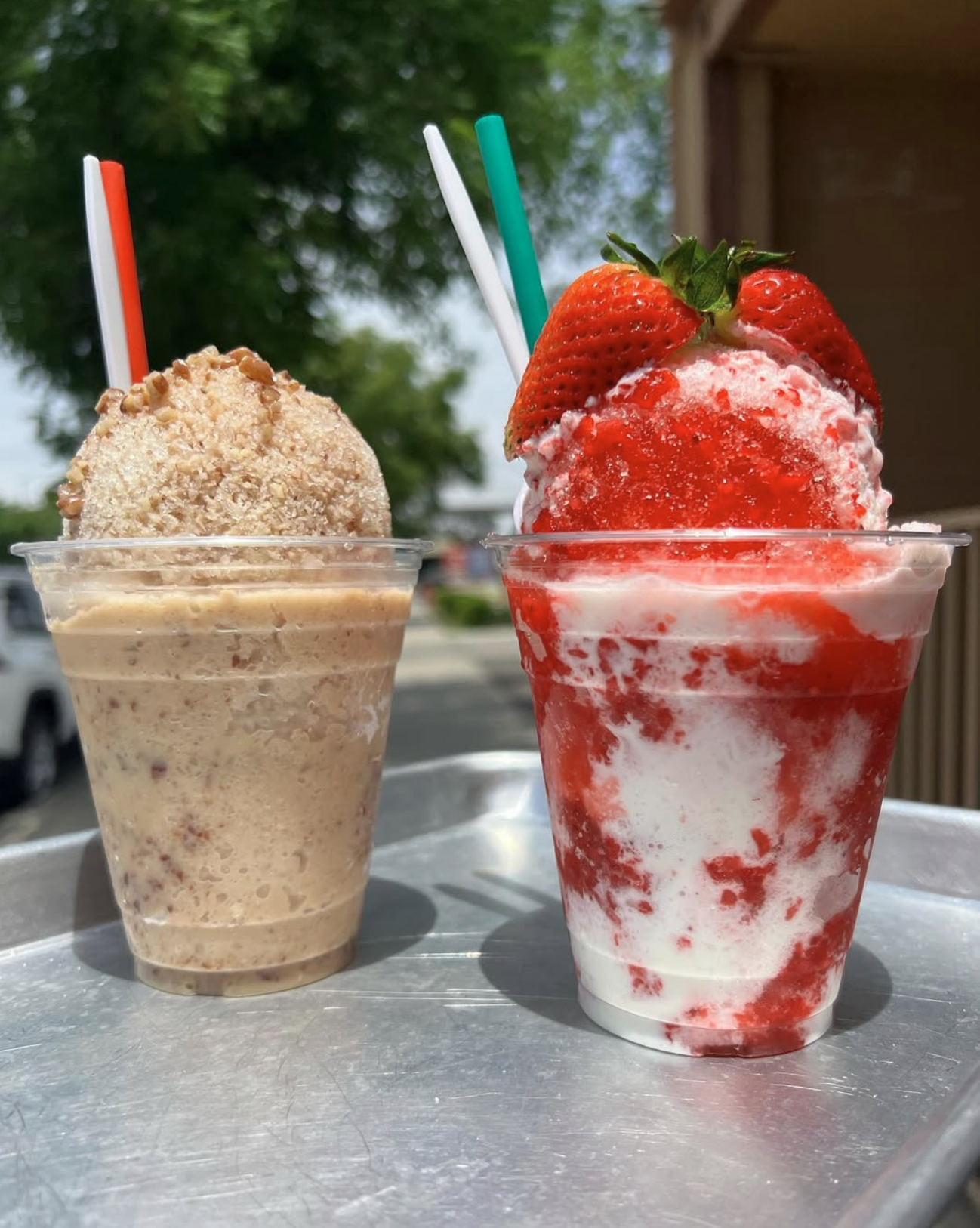 Two milkshake drinks in clear plastic cups on a metal tray, one with a beige nut and cream flavor topped with chopped nuts, the other with a red and white strawberry flavor topped with fresh strawberries and green leaves, outdoors with green trees and a building in the background.
