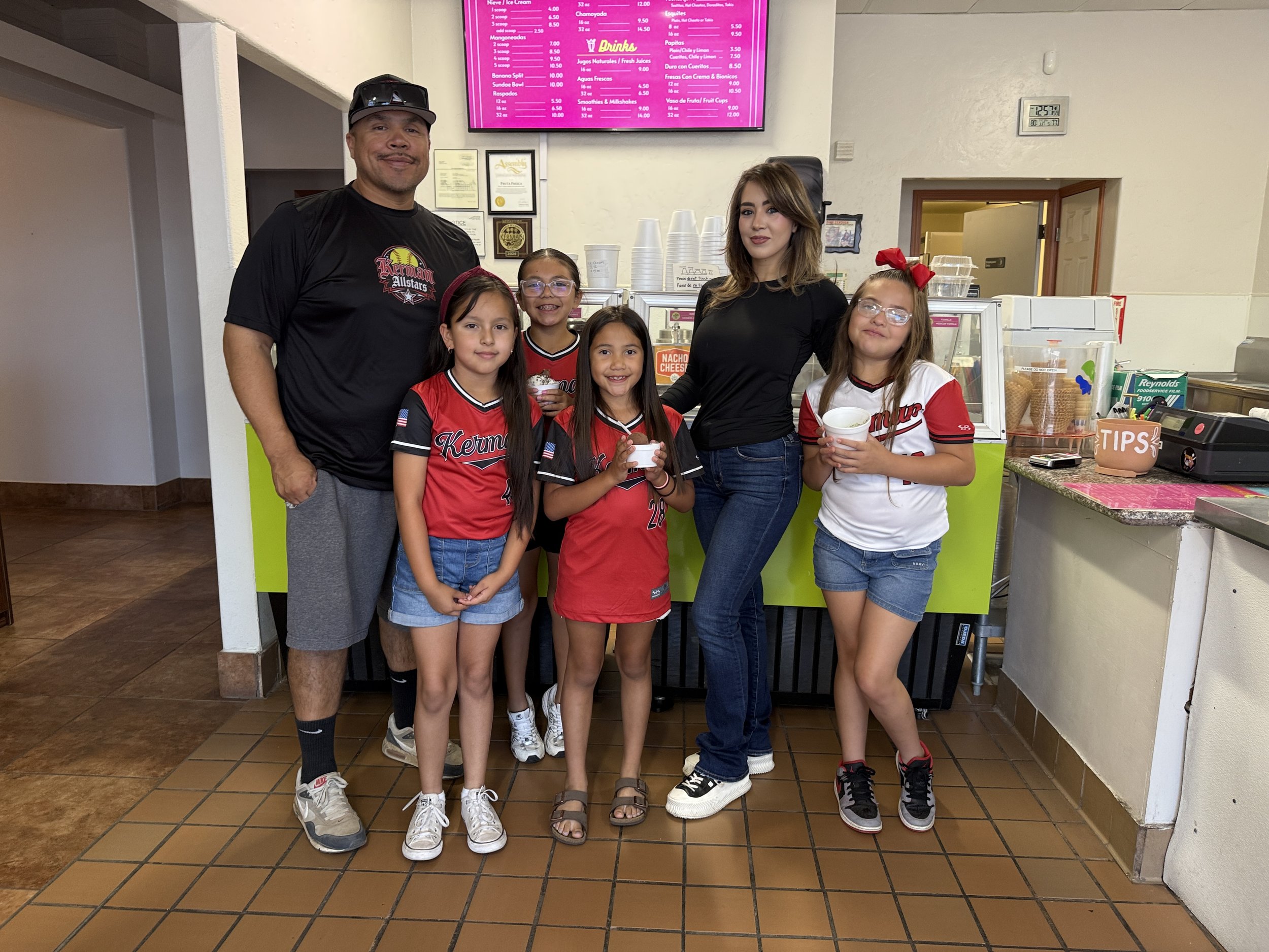 A man, woman, and three young girls are standing inside a restaurant, smiling at the camera. The girls are wearing red and white sports jerseys, and the woman is dressed in a black top and jeans. The man is wearing a black T-shirt and gray shorts. In the background, there is a bright pink menu board, some certificates on the wall, and a counter with stacks of cups and ice cream cones.