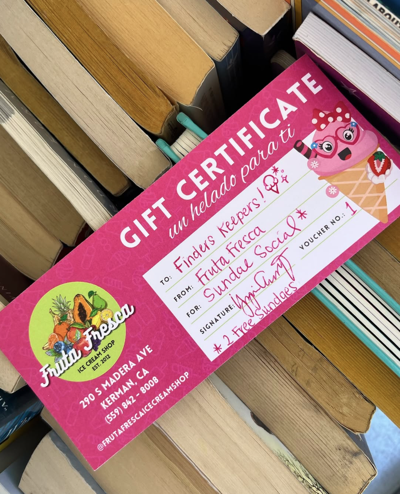 A gift certificate from Fruta Fresa ice cream shop, pink in color, featuring a cartoon ice cream cone with glasses and a bow, with handwritten details including recipient as Finder Keepers, from Fruta Fresa, for sundae social, signed by the owner, and voucher number 1. The certificate is placed on a wooden surface among other books and boxes.