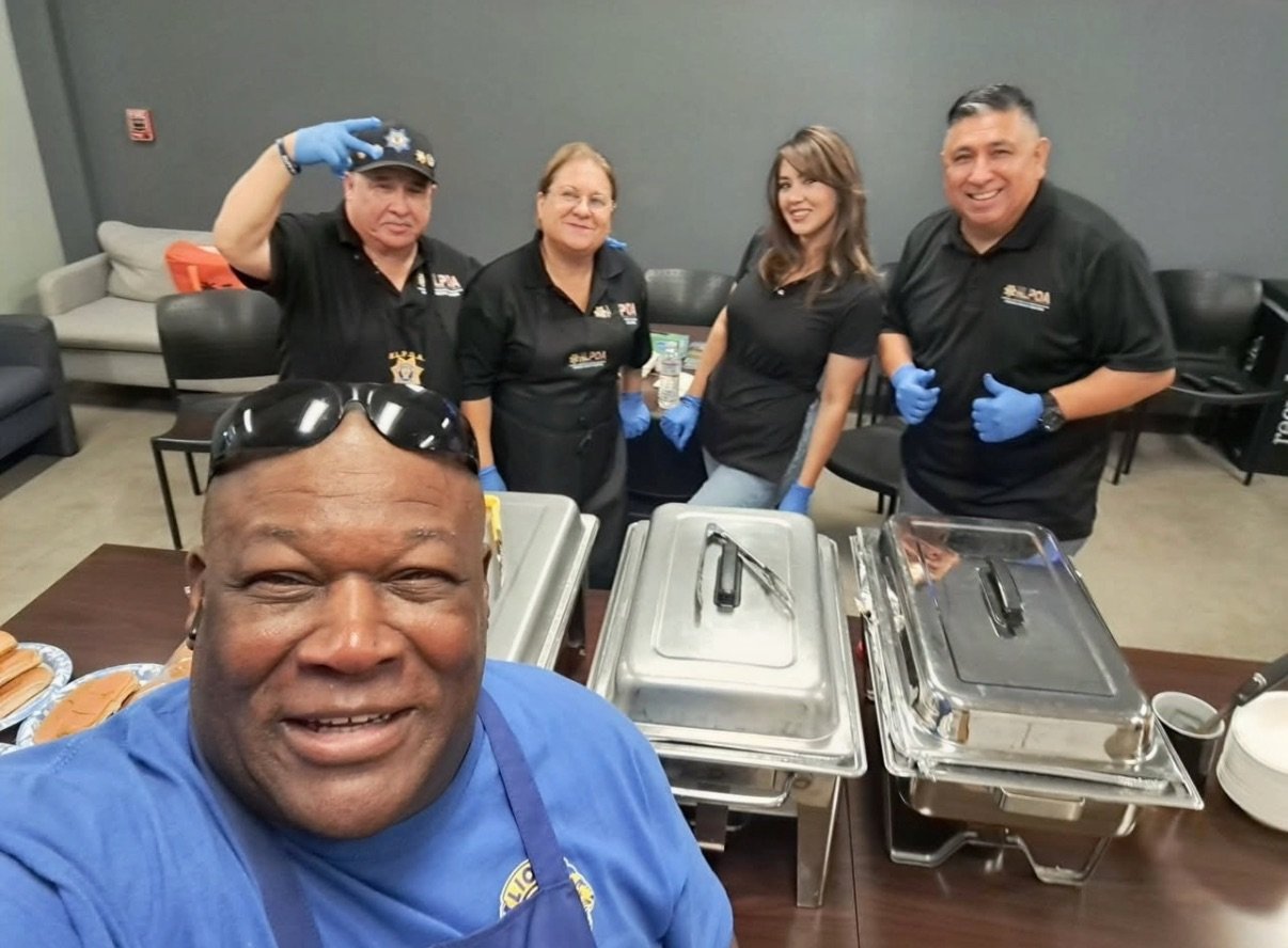 Yazmine Enriquez, 2026 Kerman Mayor Candidate and a Group of five people in black uniforms and blue gloves standing behind a table with food trays, taking a selfie, with a man in a blue shirt in the foreground wearing sunglasses.
