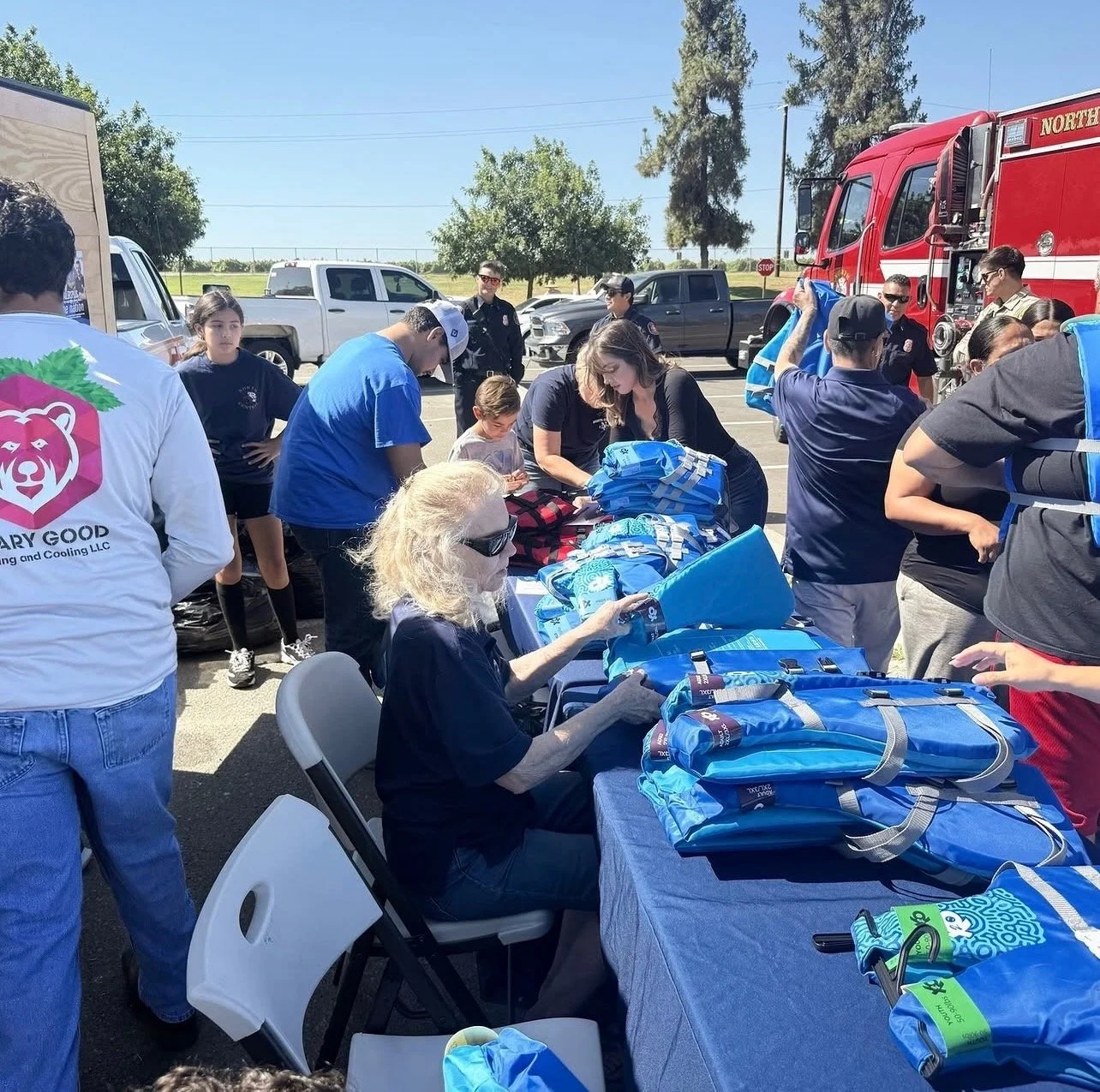 People gathered around a table with backpacks and supplies outdoors, with fire trucks and vehicles in the background, likely at a community event or disaster preparedness activity.