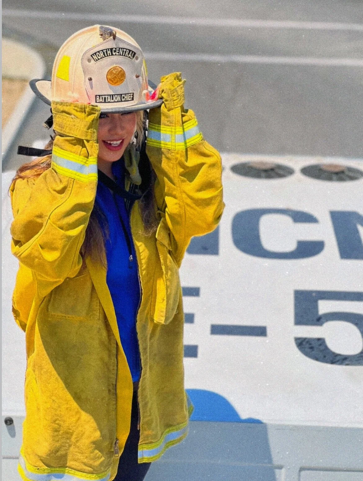 A woman in firefighter gear, including a yellow jacket and helmet, standing on an aircraft carrier or military vessel, adjusting her helmet.