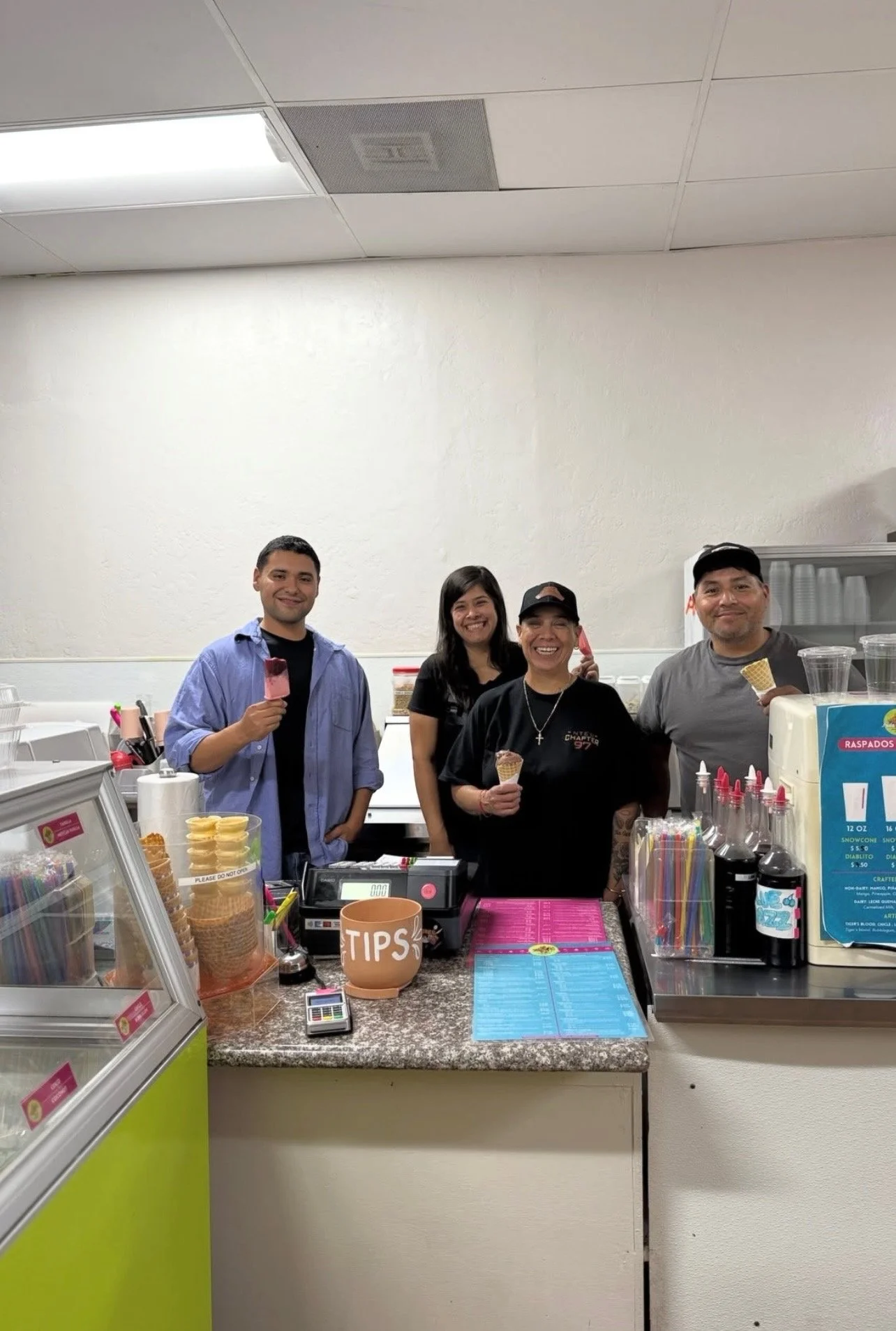 Four people smiling and holding ice cream cones behind a counter in an ice cream shop.
