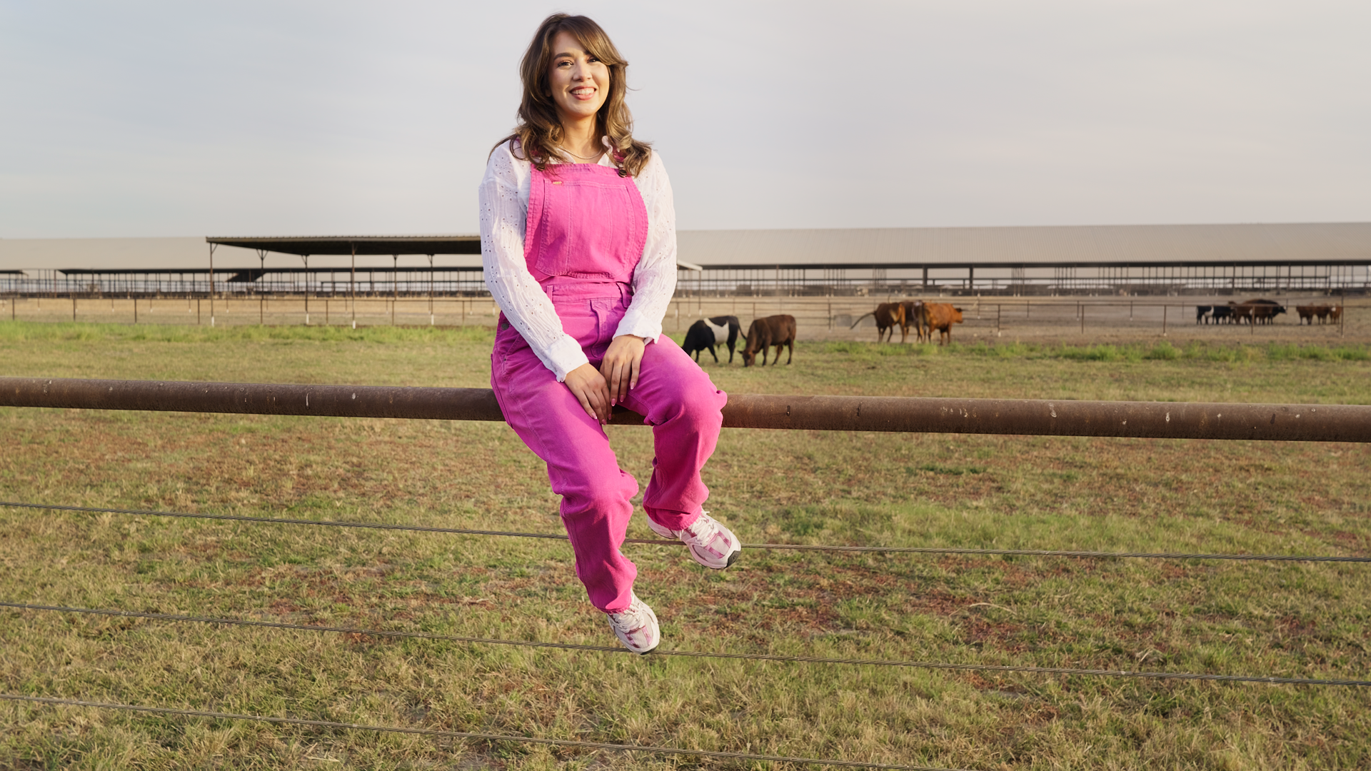 Yazmine Enriquez, 2026 Kerman Mayor Candidate sitting on a fence at a farm, smiling and winking, with cows and a barn in the background.