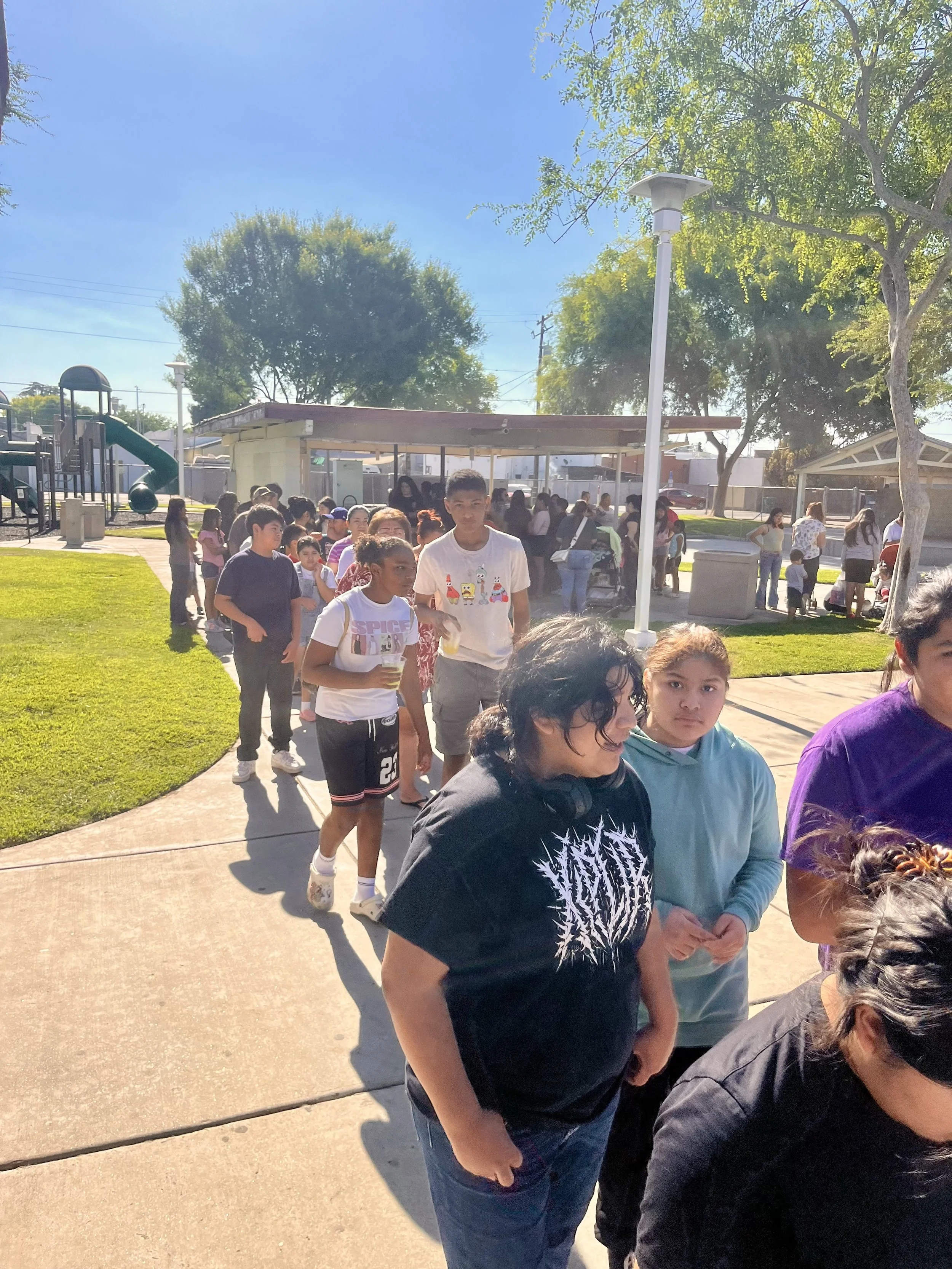 People standing and walking in line outdoors on a sunny day, with trees, a playground, and a public space in the background.