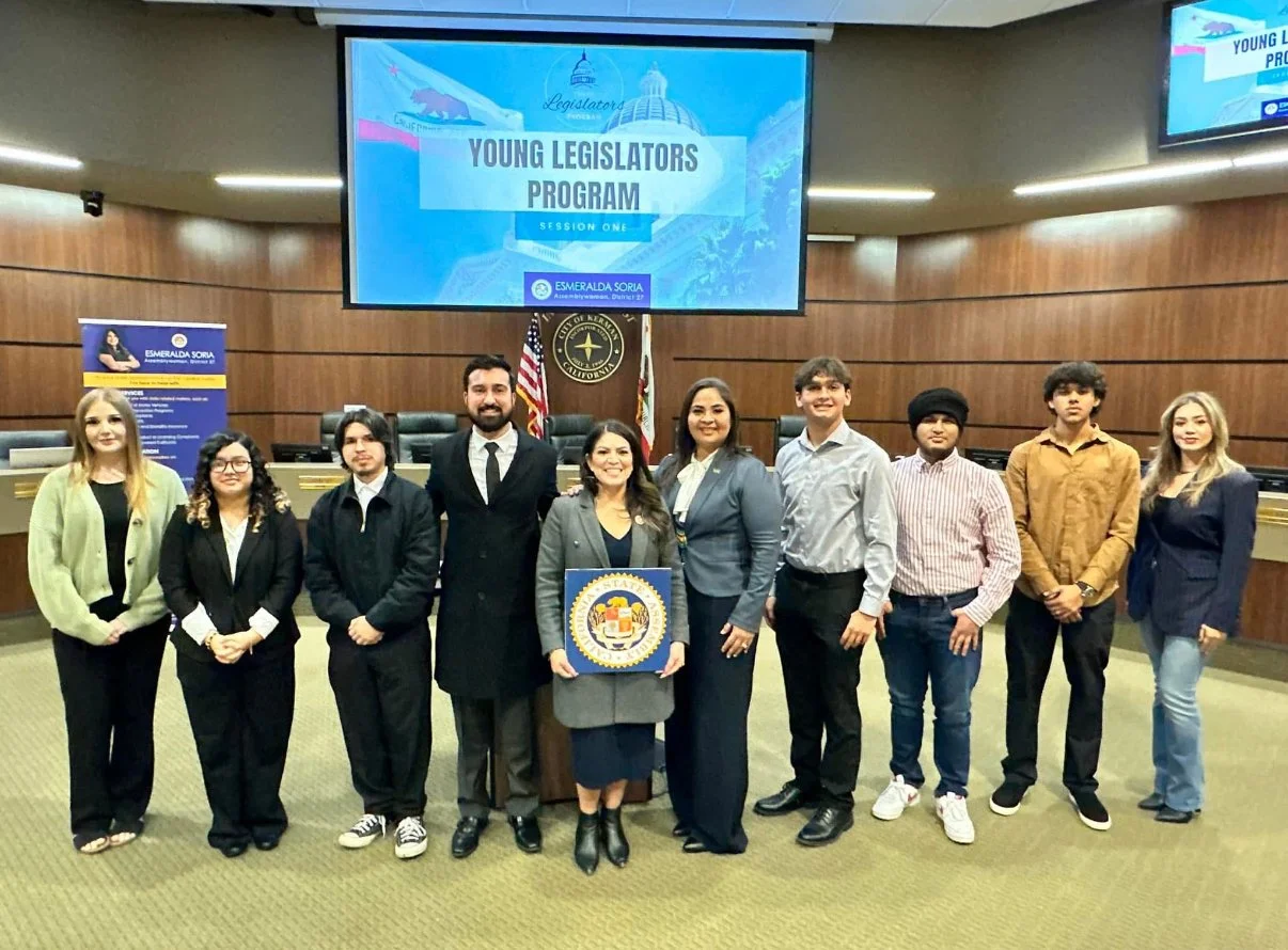 Group of young diverse people standing in a room with a large screen overhead that reads 'Young Legislators Program, Session One' and features the California Legislature logo. They are posing for a photo, some holding a seal of California or a plaque, with flags in the background.
