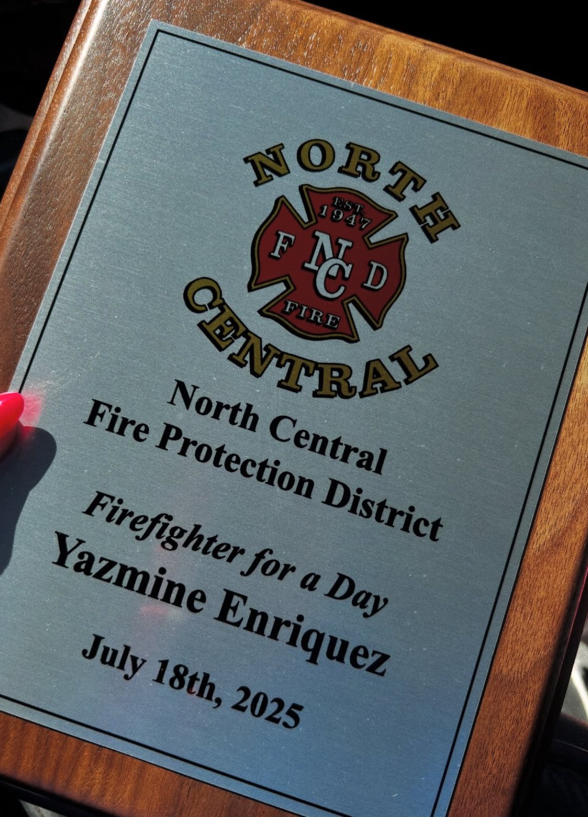 A plaque for the North Central Fire Protection District, featuring the district's emblem, which includes a red fire cross with the letters FC D, and details about an event honoring firefighter Yazmine Enriquez on July 18th, 2025.
