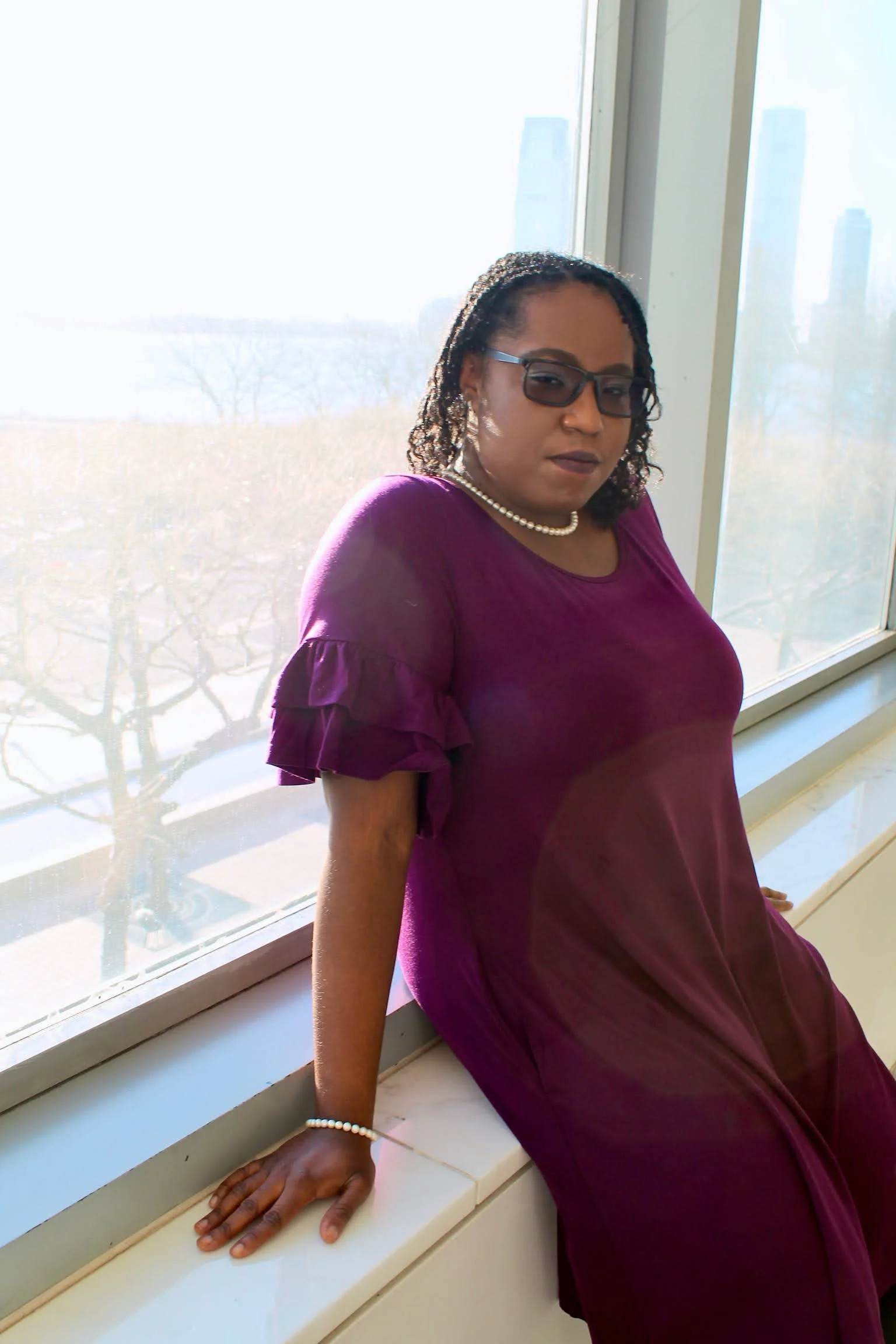 A woman with dark curly hair, wearing glasses, a pearl necklace, and a purple dress with ruffled sleeves, sitting on a white window sill near large windows with a cityscape and trees outside.