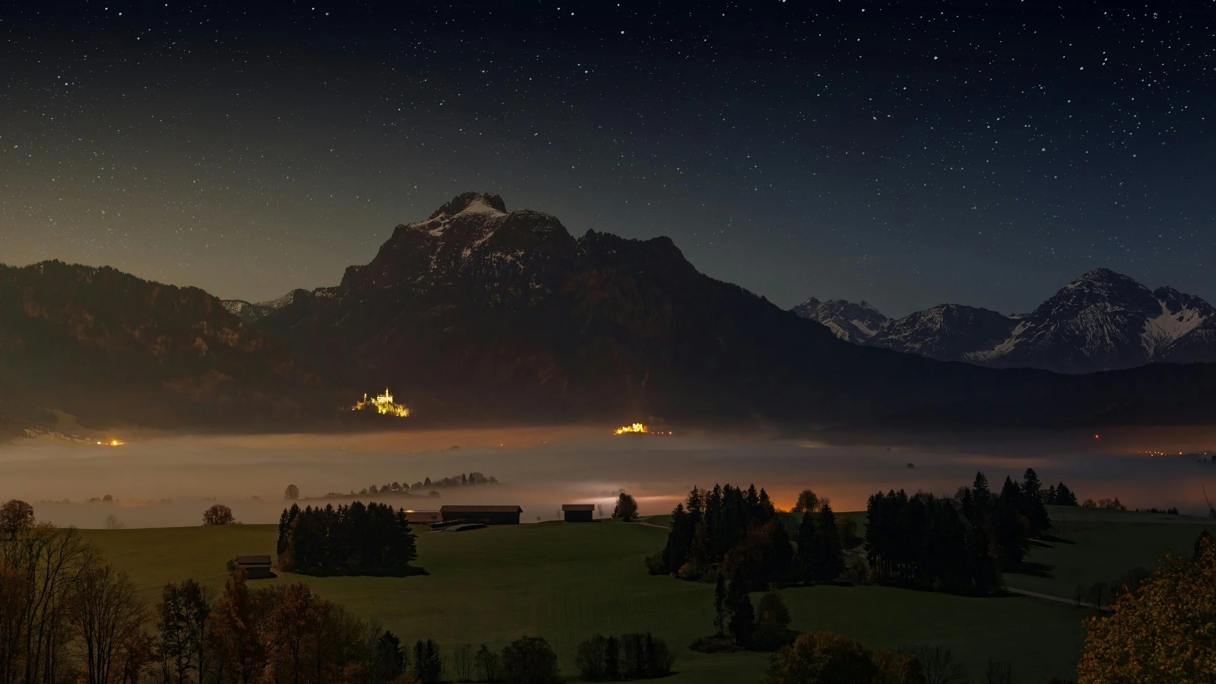 Nighttime landscape featuring mountains, a starry sky, fog in the valley, illuminated buildings, and trees in the foreground.