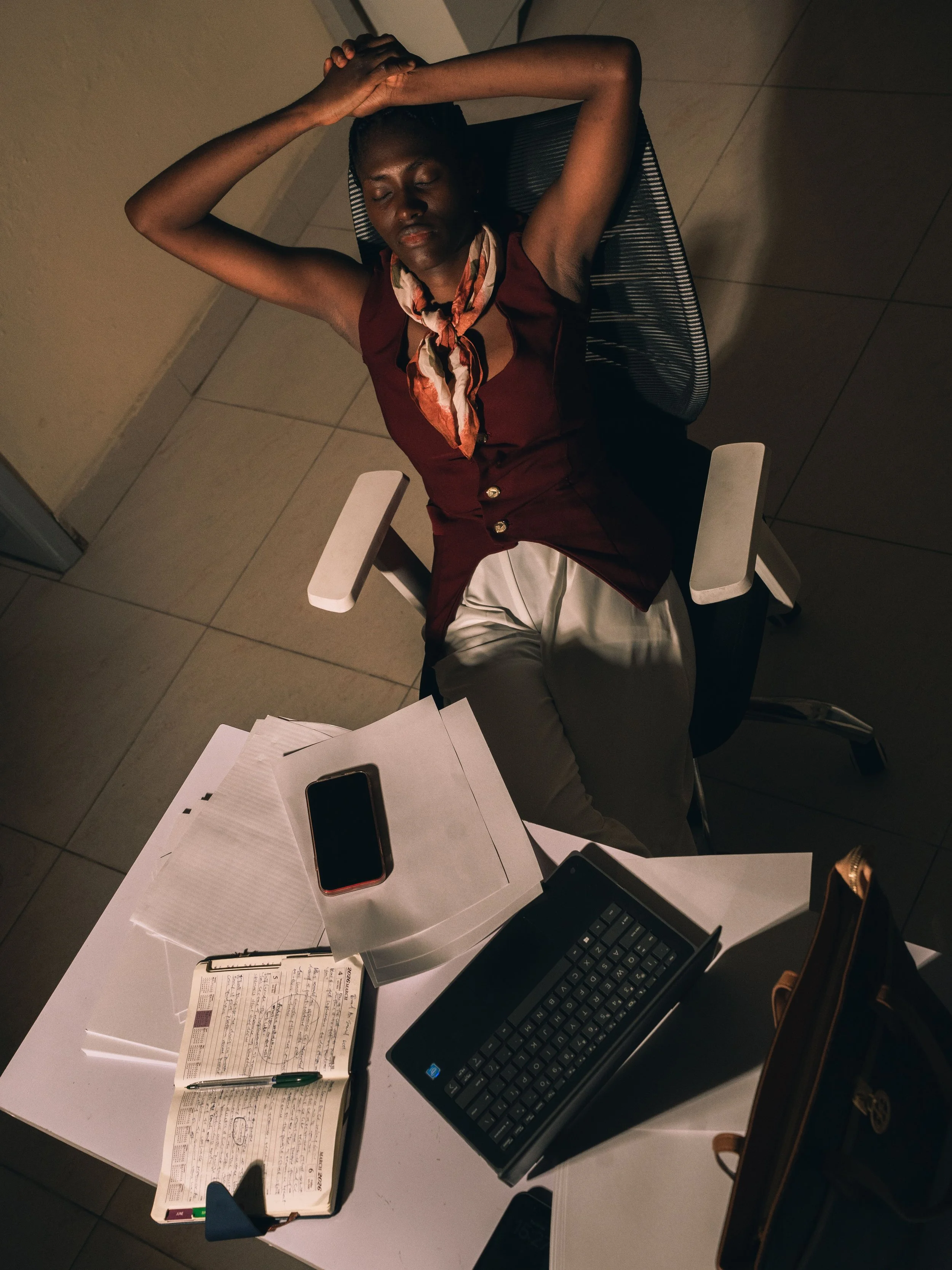 A woman sitting at a desk with her eyes closed and hands on her head, appearing stressed or tired. The desk contains papers, a pen, a notebook, a computer keyboard, and a smartphone. She is wearing a sleeveless maroon top and a scarf around her neck. The setting appears to be an office or a workspace.