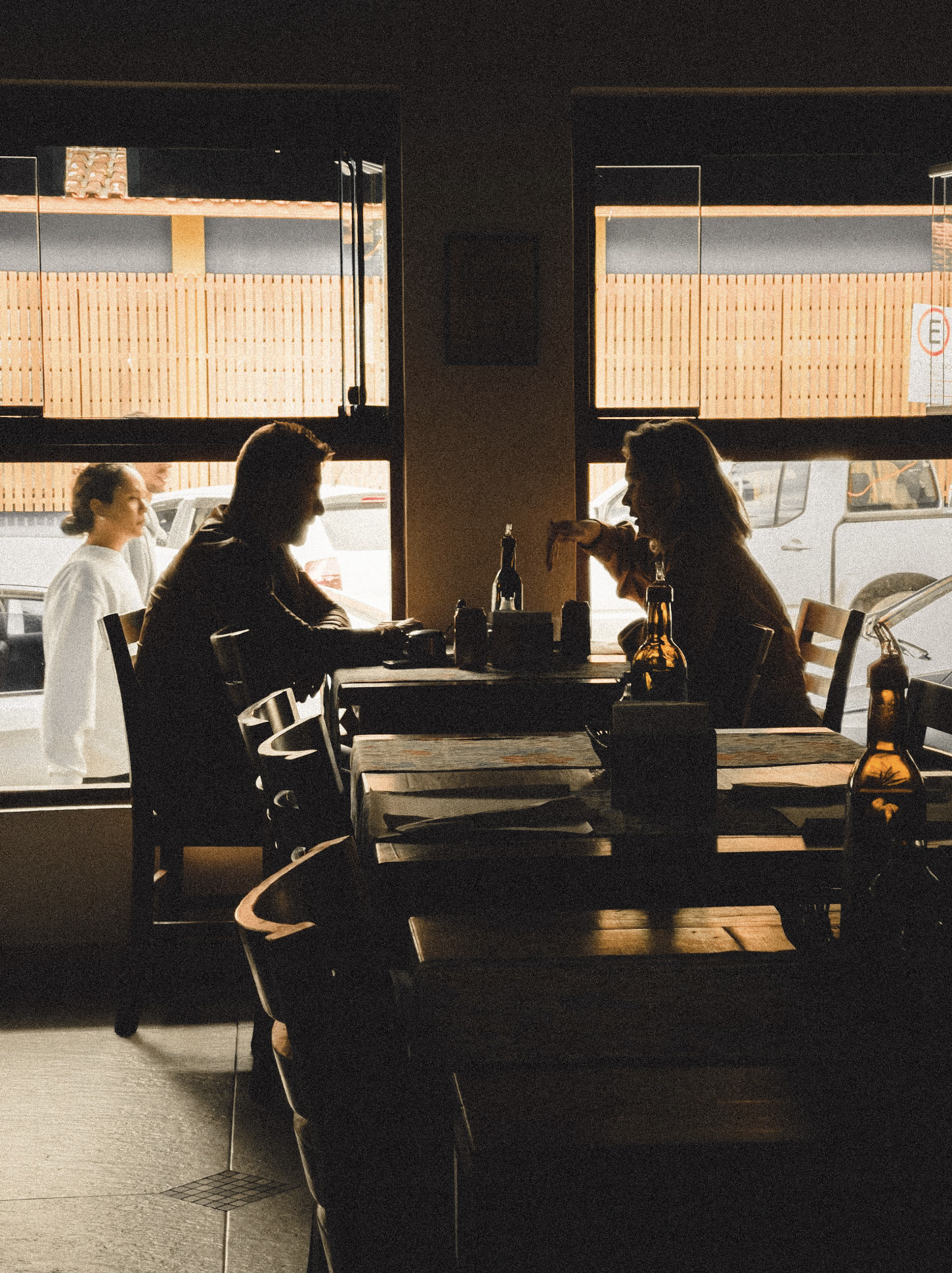 Inside a restaurant or cafe with three people sitting at a table, seen through two large windows. Outside views include parked cars and wooden panels, with sunlight illuminating the scene.