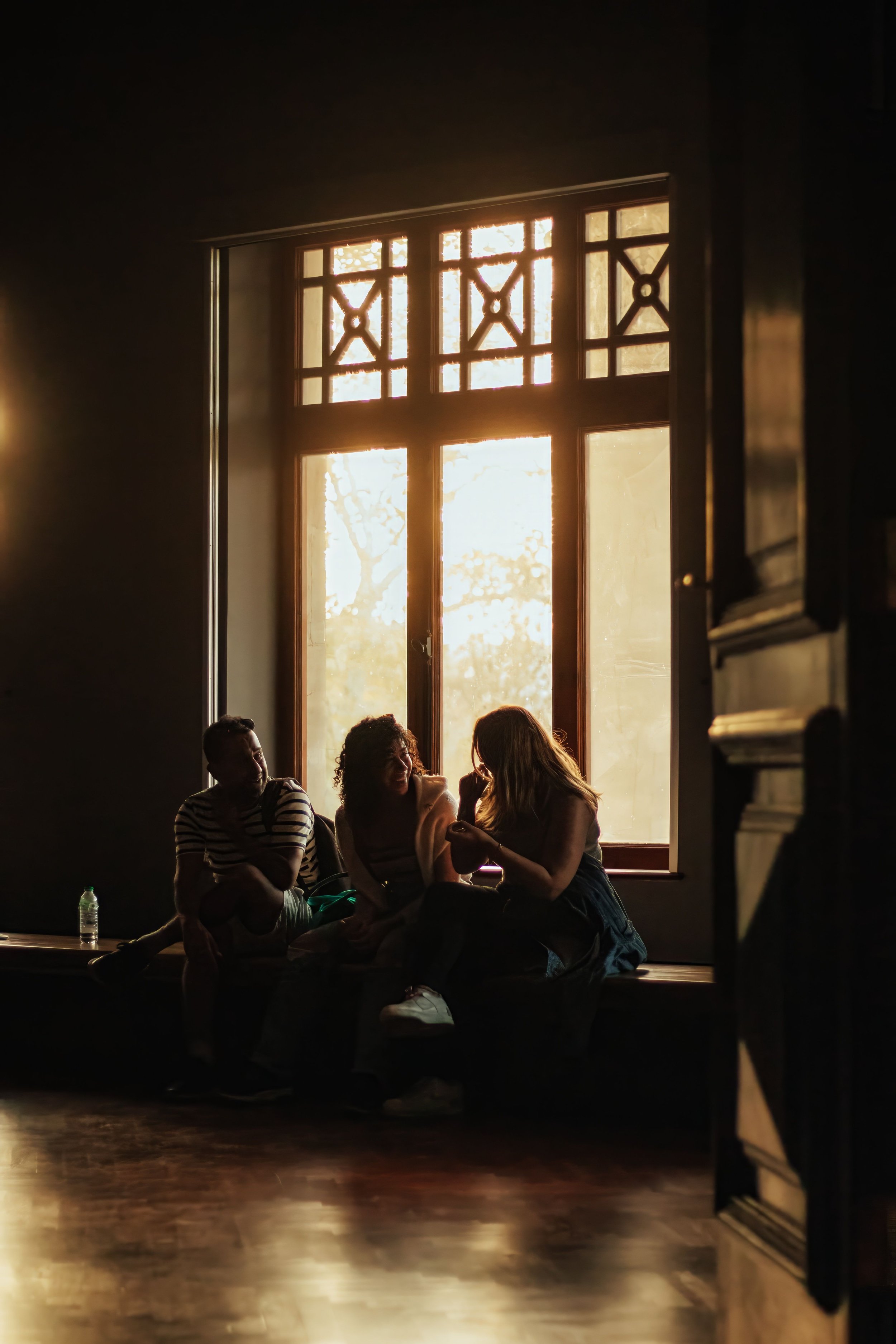 People sitting and talking by a large window with decorative grid patterns, sunlight streaming in, and dim indoor lighting.