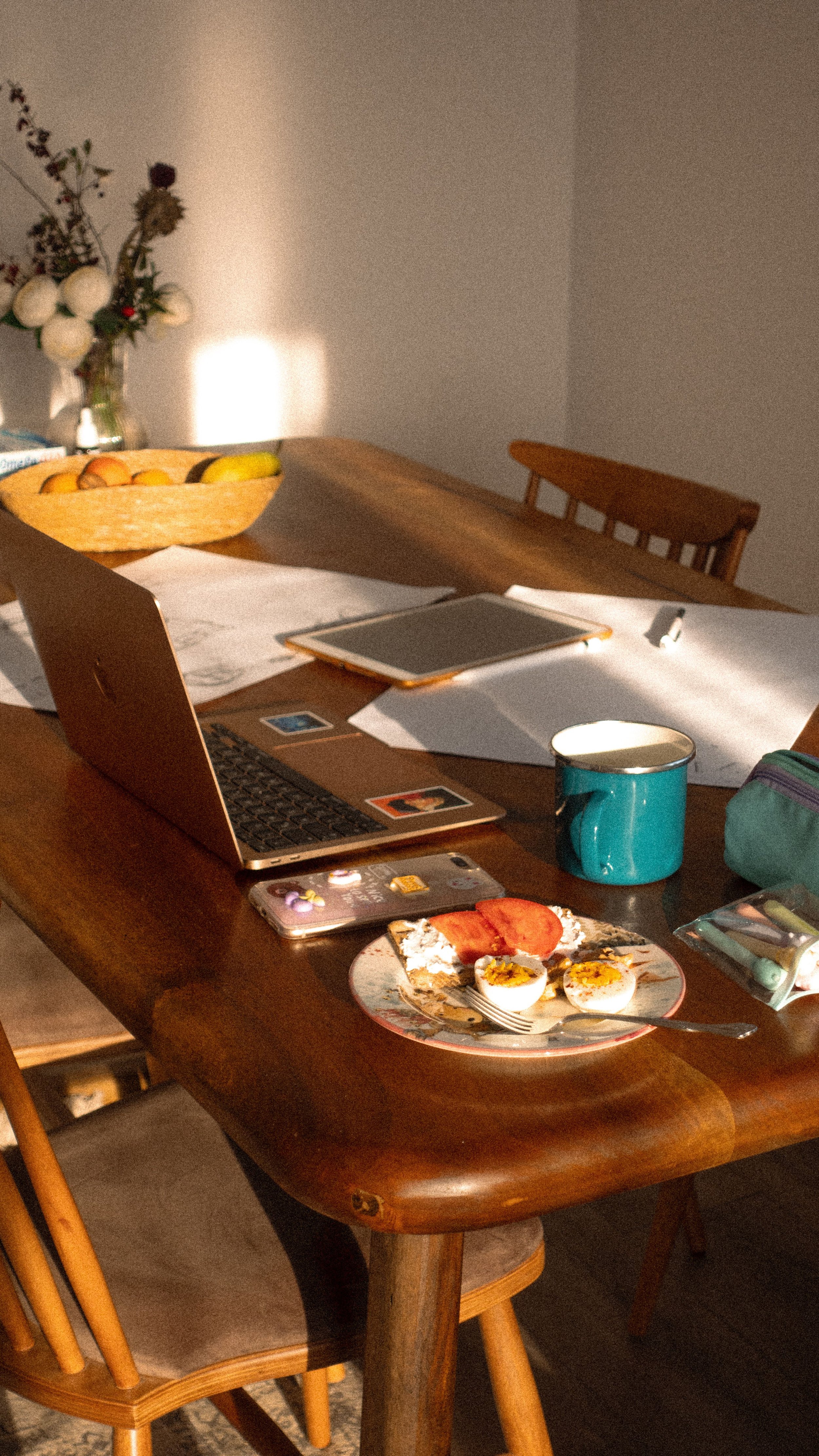 A dining table with open laptop, papers, a tablet, a pen, a mug, and a plate with boiled eggs, tomato, and bread. In the background, there is a flower vase with white flowers and a fruit bowl with bananas and oranges, with sunlight casting shadows on the wall.