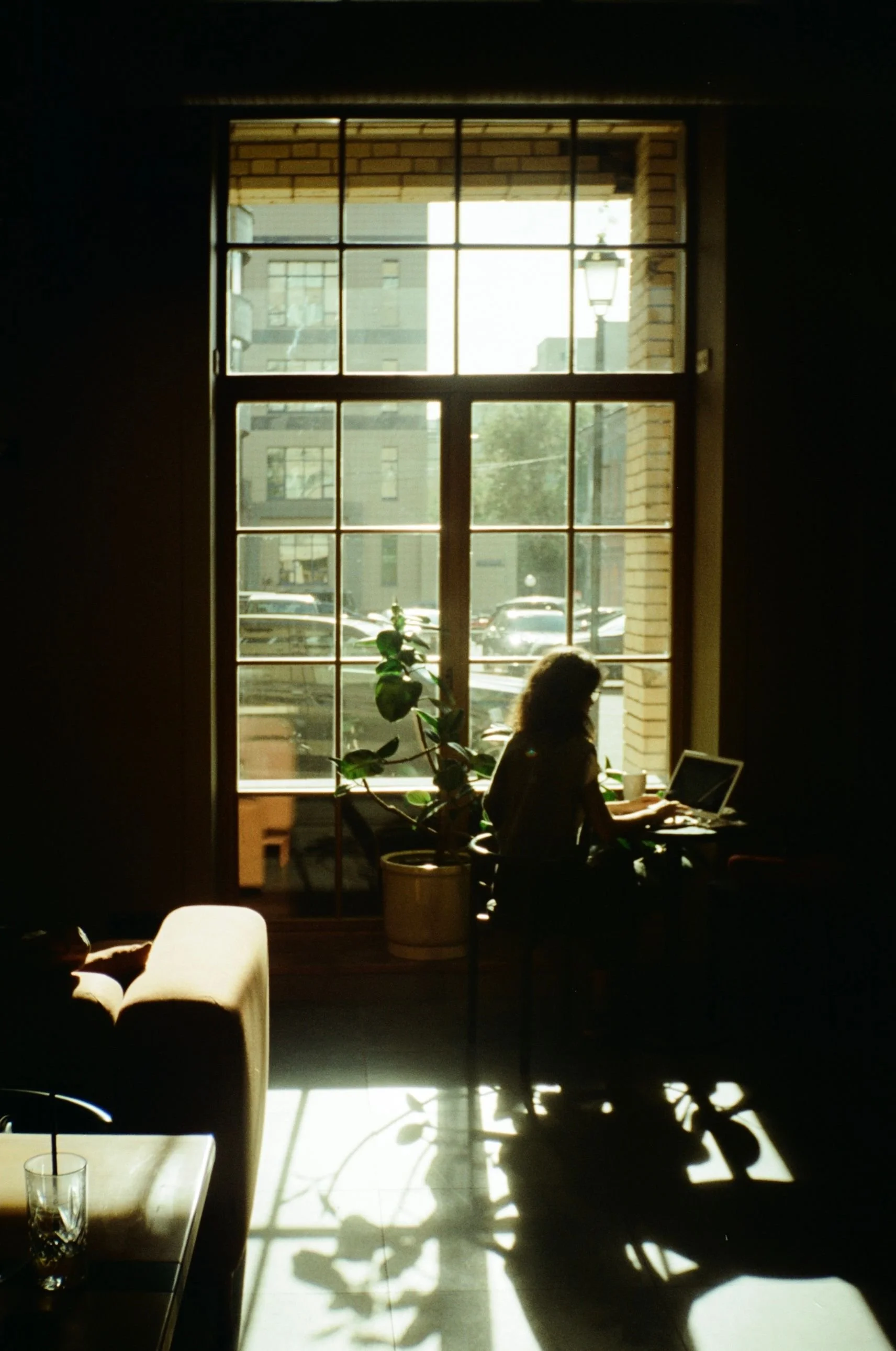 A person sitting at a desk working on a laptop near a large window with sunlight streaming in, casting shadows on the floor and furniture.