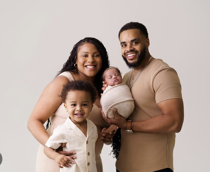 Family portrait of five, including Shanbria Oglesby, Founder and Licensed Professional Counselor, man, a young boy, and a newborn baby wrapped in a blanket, standing close together and smiling against a plain background.