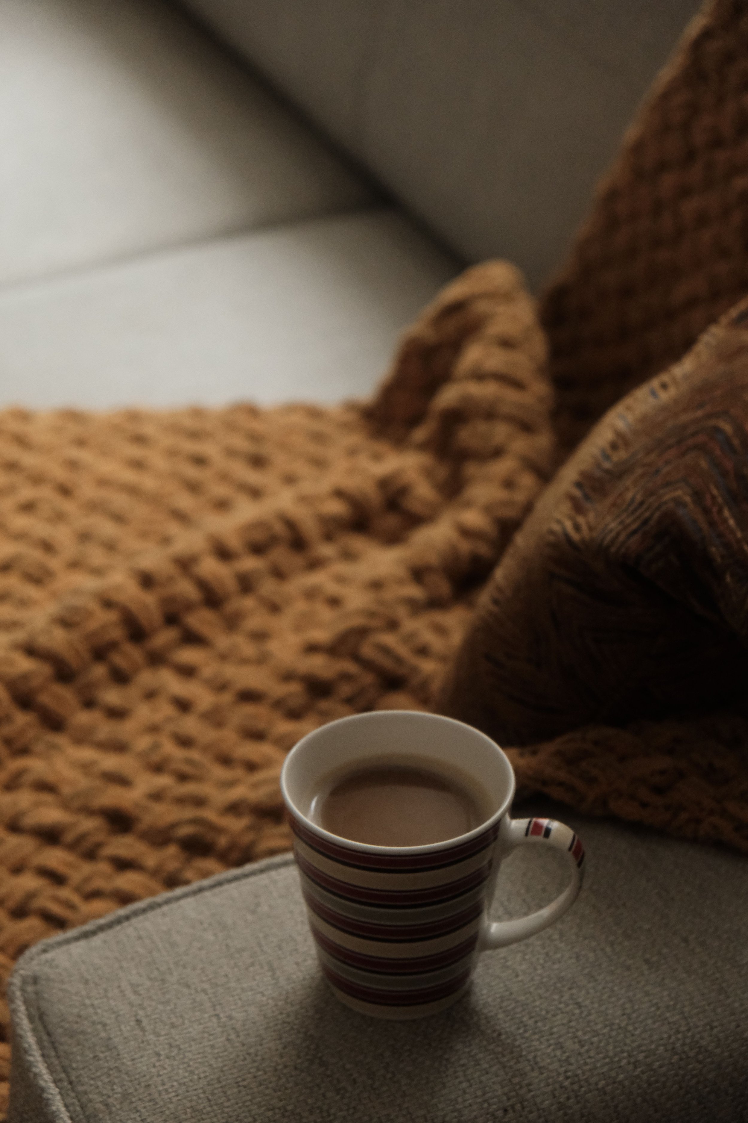 A mug of coffee or tea with red and white stripes is placed on a small white table. In the background, there is a brown textured blanket and a patterned cushion on a couch. Part of a white wall and ceiling are also visible.