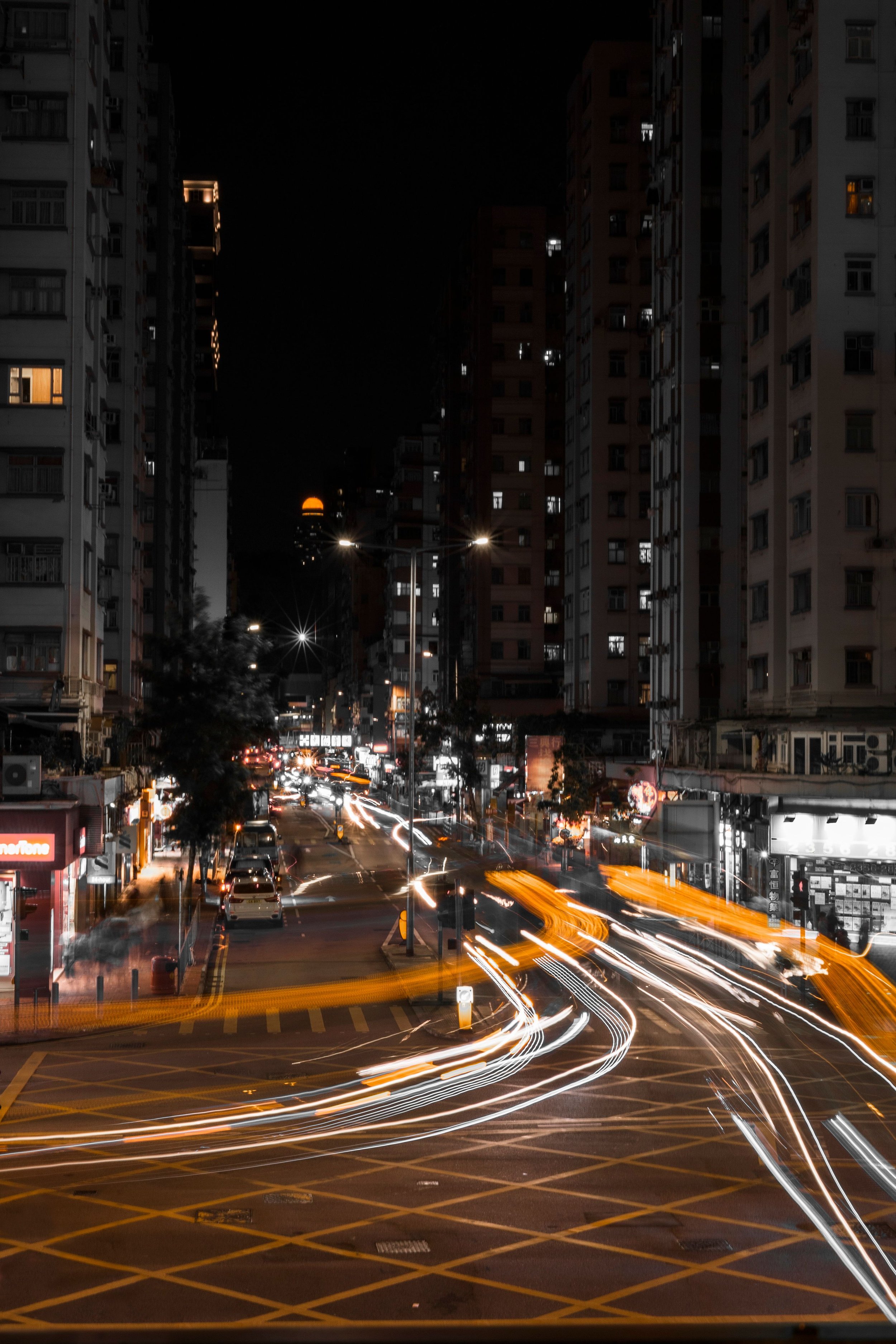 Nighttime city street scene with tall apartment buildings, cars, and light trails from moving vehicles, with some buildings illuminated.