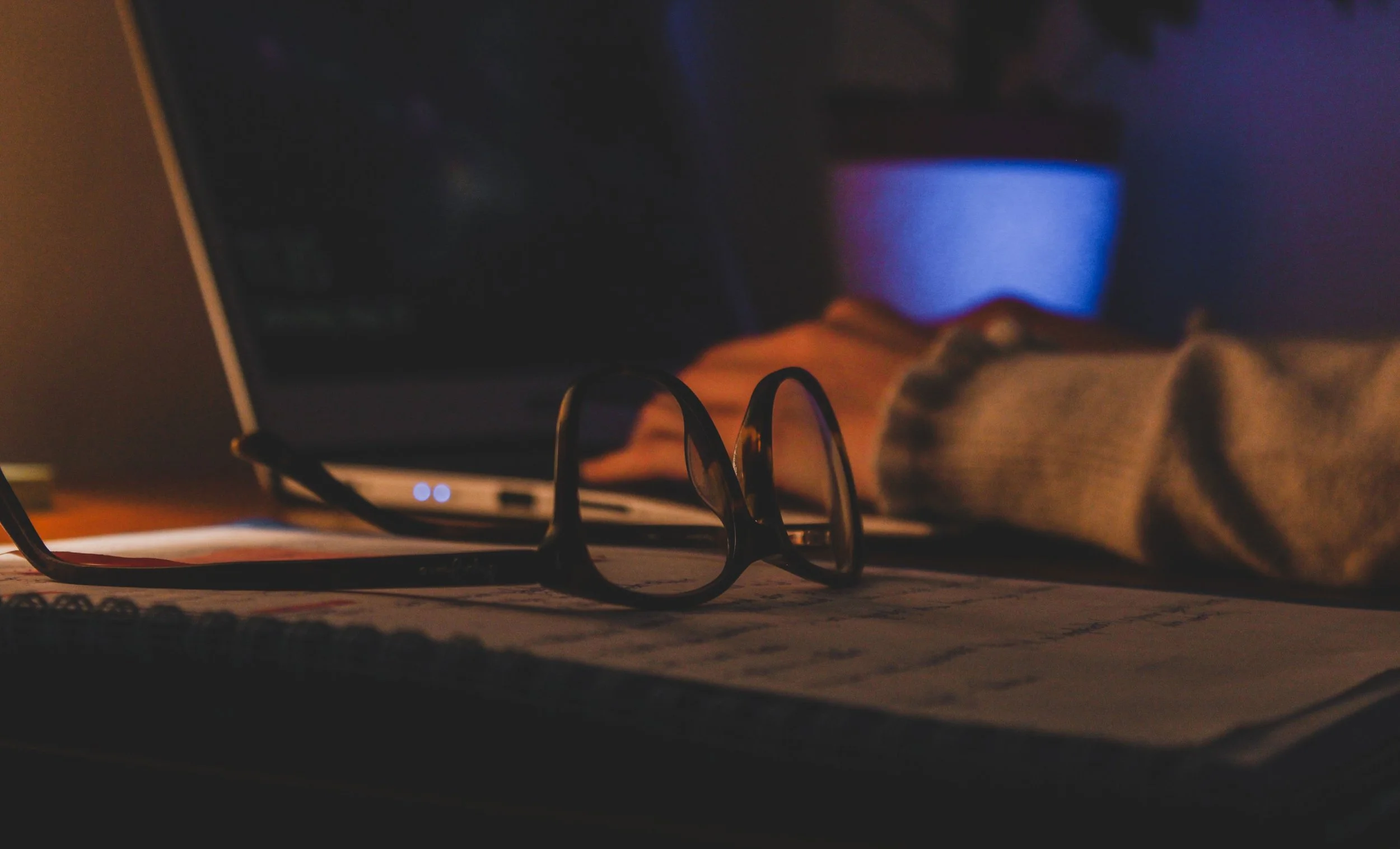 A pair of eyeglasses resting on a table with an open notebook underneath, in front of a computer screen showing a blue-toned image, with a lit LED indicator on the computer and a blurred background.