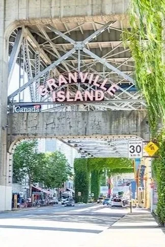 View of Granville Island sign under a bridge over a city street with trees and buildings.