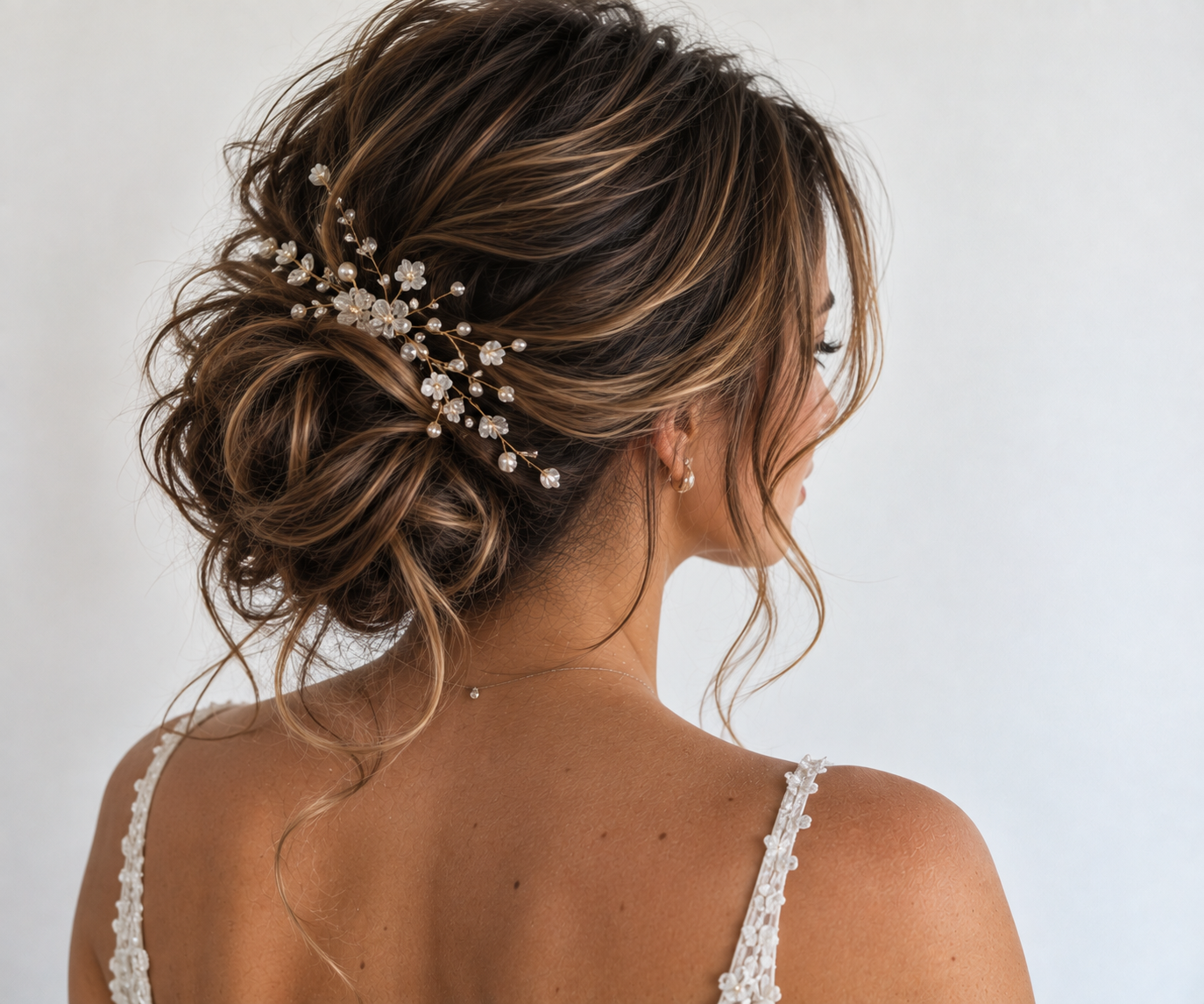 A woman with styled brunette hair with blonde highlights, wearing a decorative hairpiece with white flowers and pearls, and a white lace dress, with a bare shoulder visible.