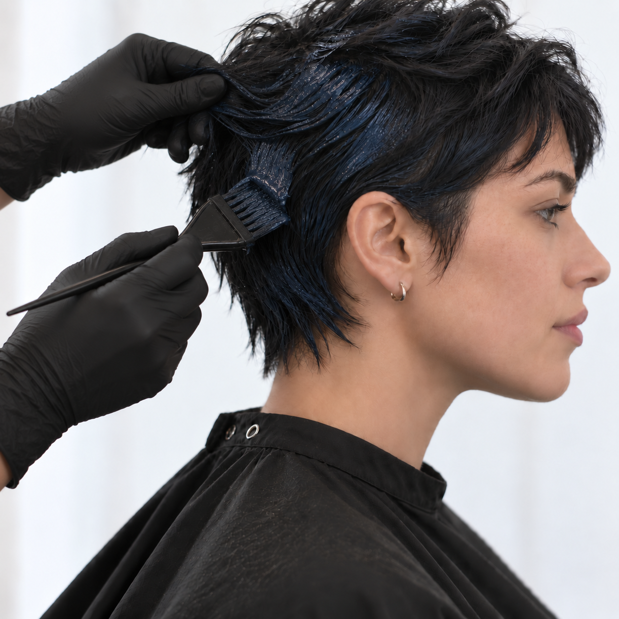 A woman with short dark hair getting her hair dyed dark blue in a salon stylist's hand, wearing a black cape.