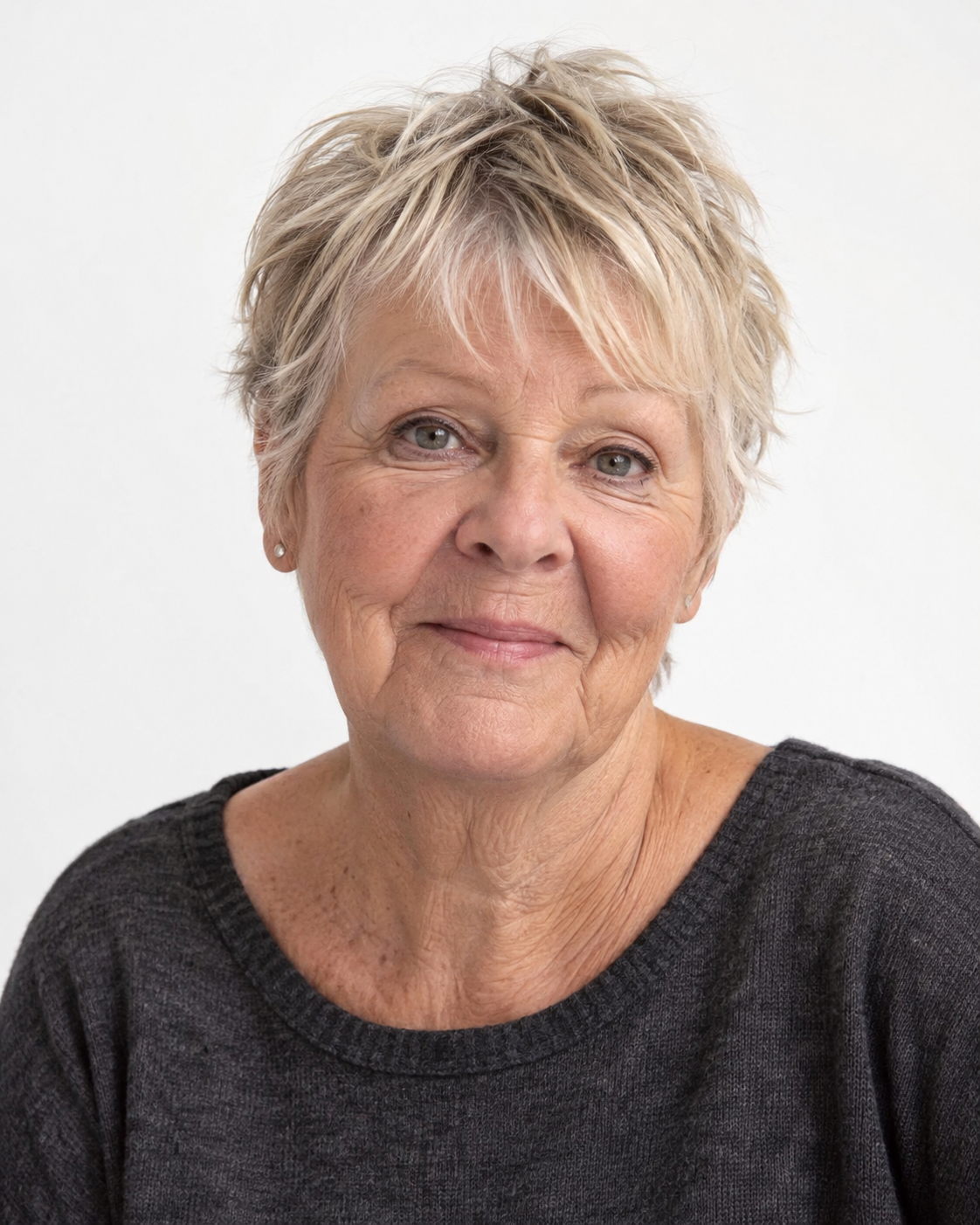 Close-up of an elderly woman with short, tousled blonde hair, wearing a black sweater, smiling softly against a plain white background.