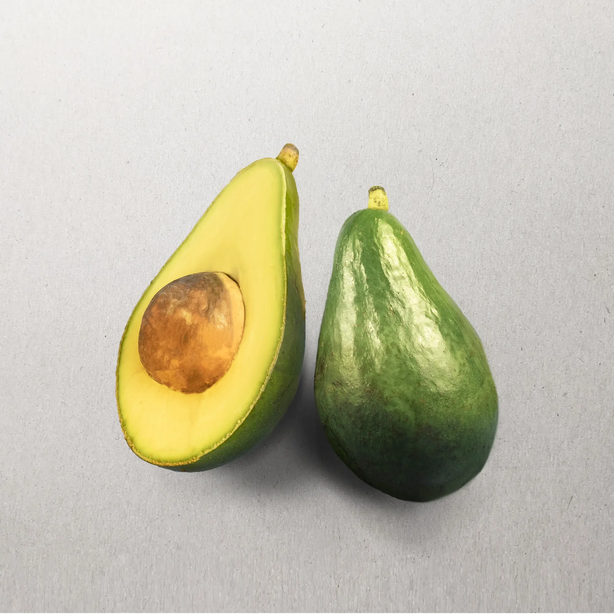 A sliced avocado and a whole avocado on a plain gray surface.