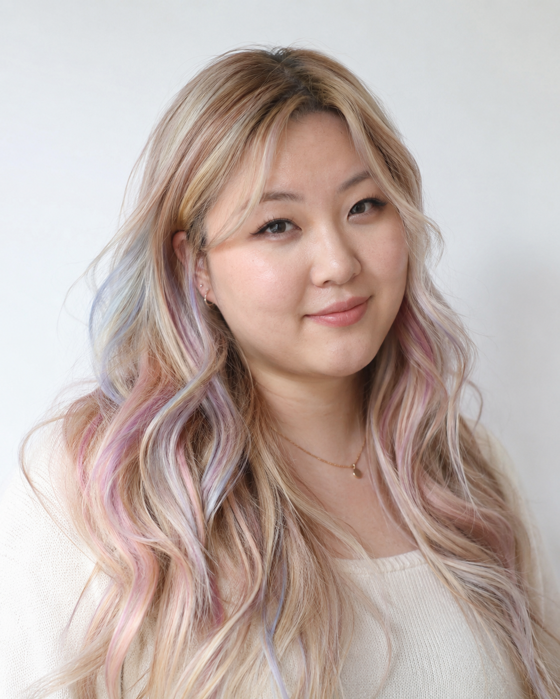 A woman with long, wavy hair dyed in pastel rainbow colors, smiling slightly, wearing a white top, with a plain light background.