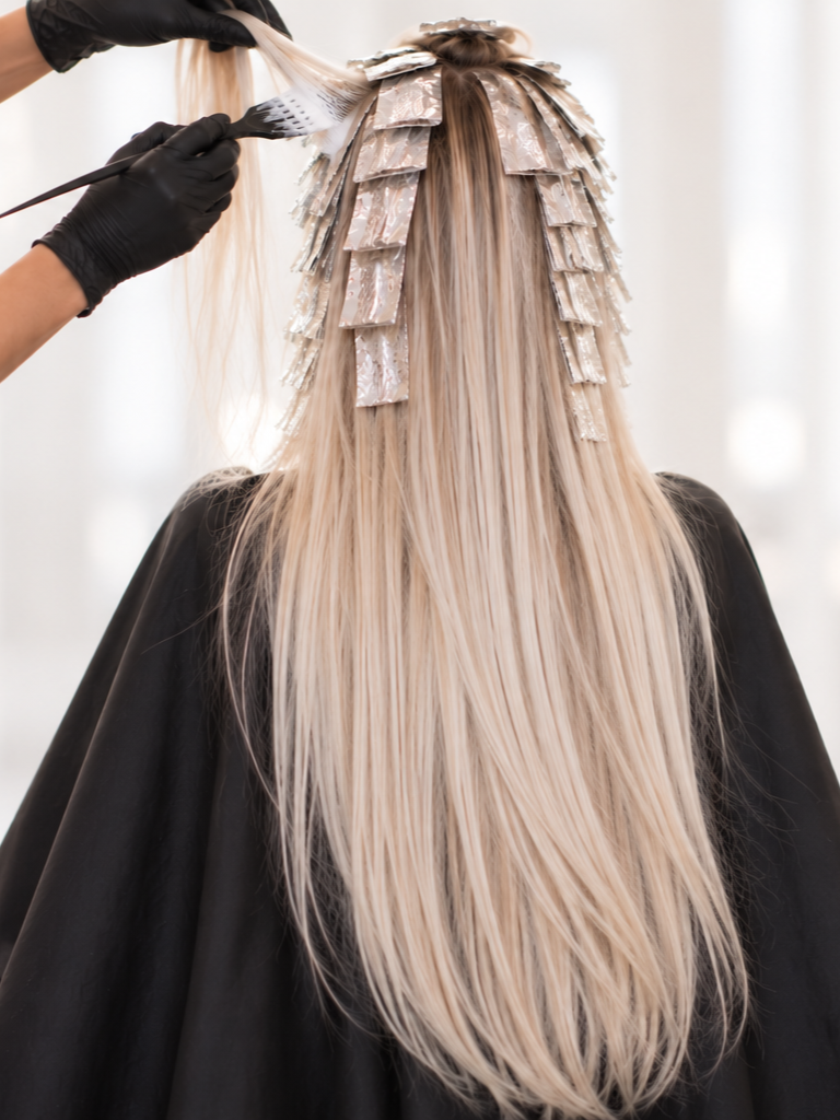 Hairdresser applying hair dye to long blonde hair, with hair held in hair color foils, in a salon setting.
