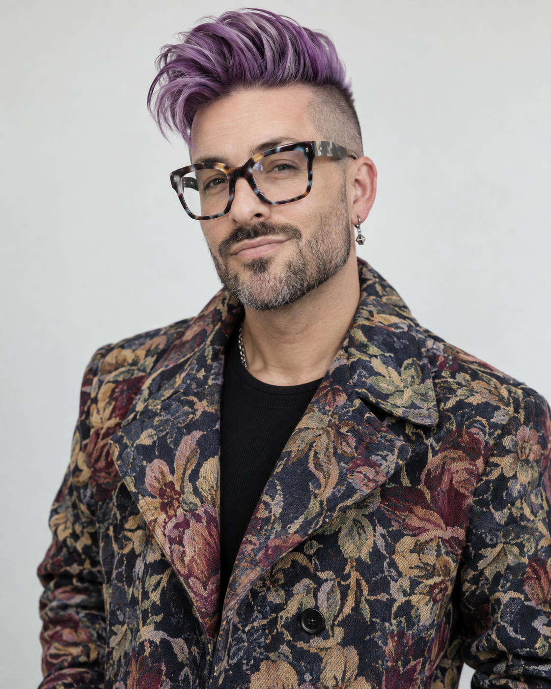 A man with purple hair, glasses, an earring, and a beard, wearing a floral patterned blazer over a black shirt, against a plain light background.