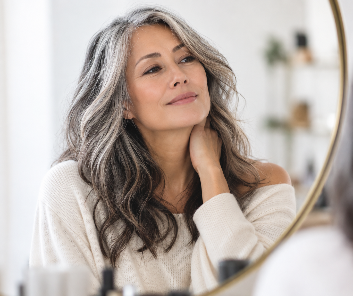 A middle-aged woman with long, wavy gray hair gazes at her reflection in a mirror, touching her neck with one hand, wearing a cream-colored sweater, with a calm and confident expression.