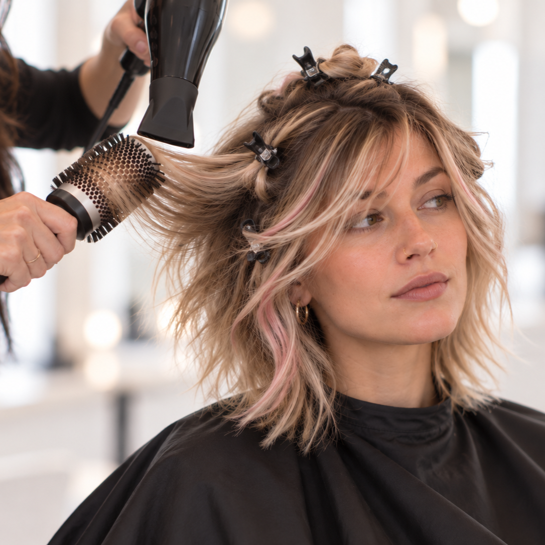 A woman with blonde, pink, and brown hair getting her hair blow-dried at a salon while clips hold her hair in place.