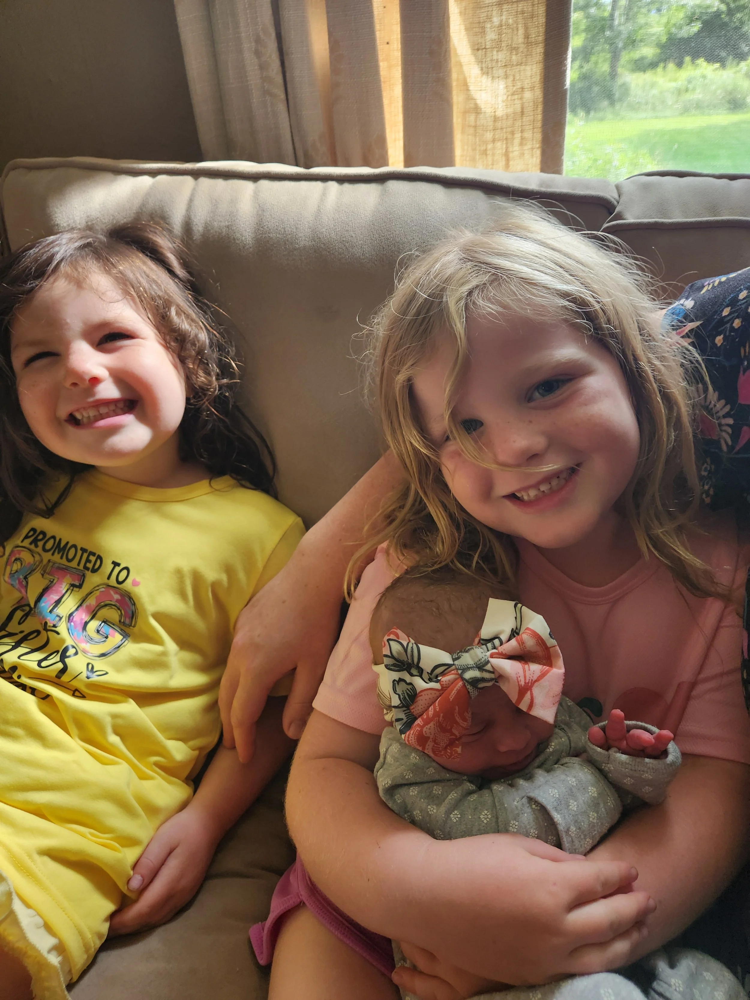 Two young girls and an infant sitting on a couch near a window, smiling and relaxing together.