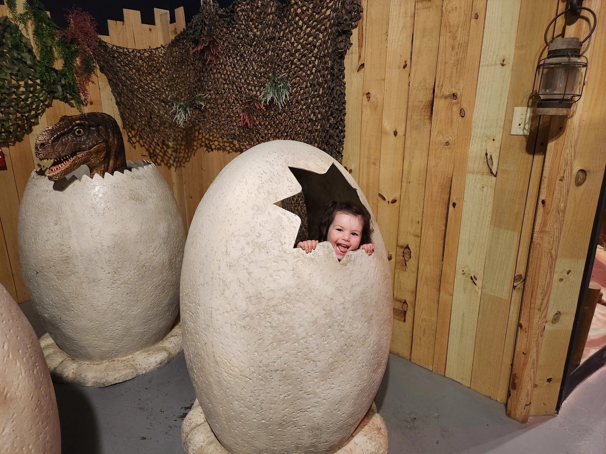 A young girl with curly hair and a big smile, peeking out of a large, cracked eggshell decoration. The scene has a wooden wall background with decorative greenery and a dinosaur head emerging from another eggshell, creating a playful Easter or spring-themed setting.