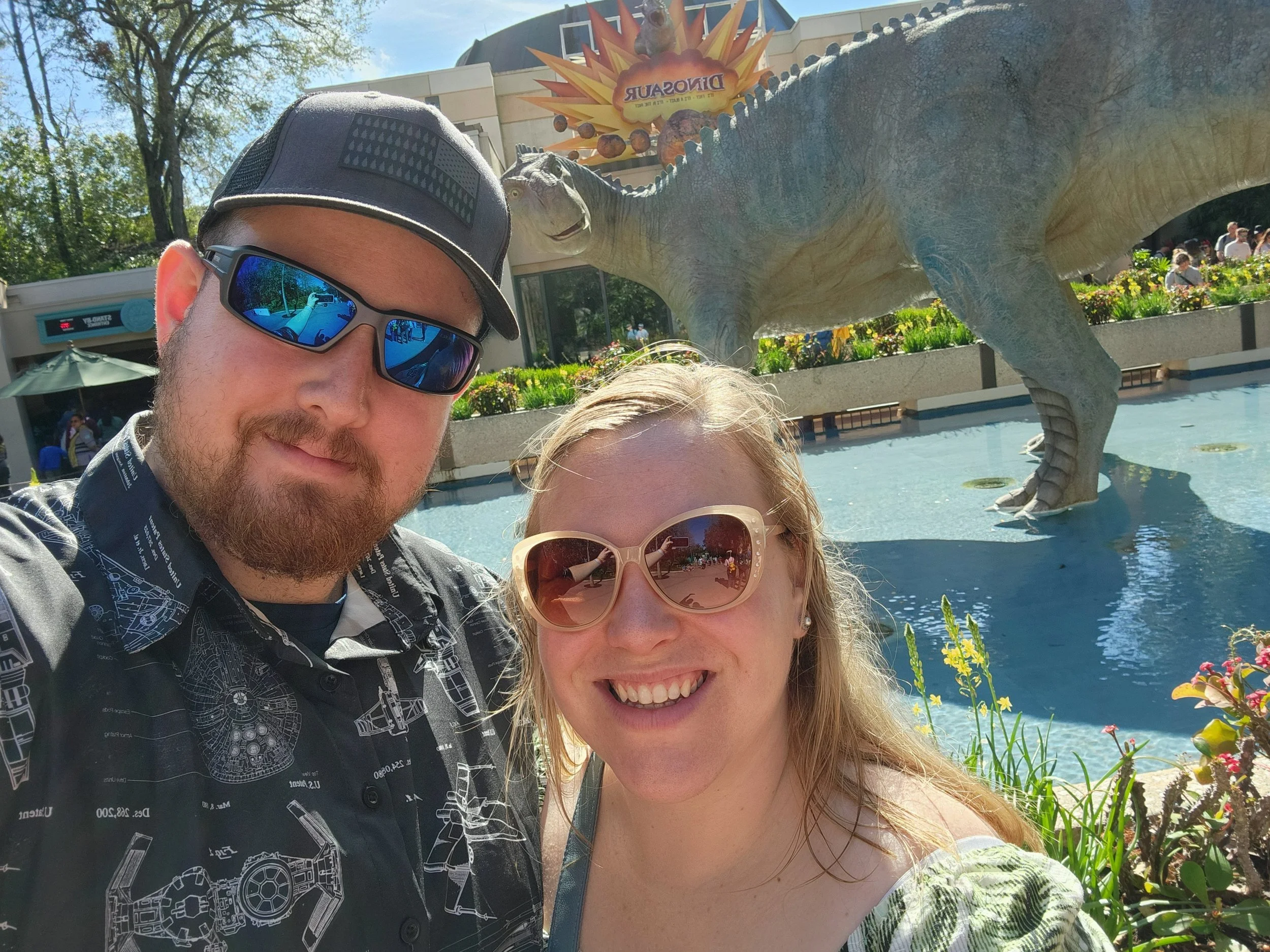 A man and woman taking a selfie in front of a dinosaur statue at an amusement park. The man is wearing sunglasses, a cap, and a dark shirt, while the woman is smiling and wearing sunglasses and a light-colored top. There are trees, flowers, and a crowd of people in the background.