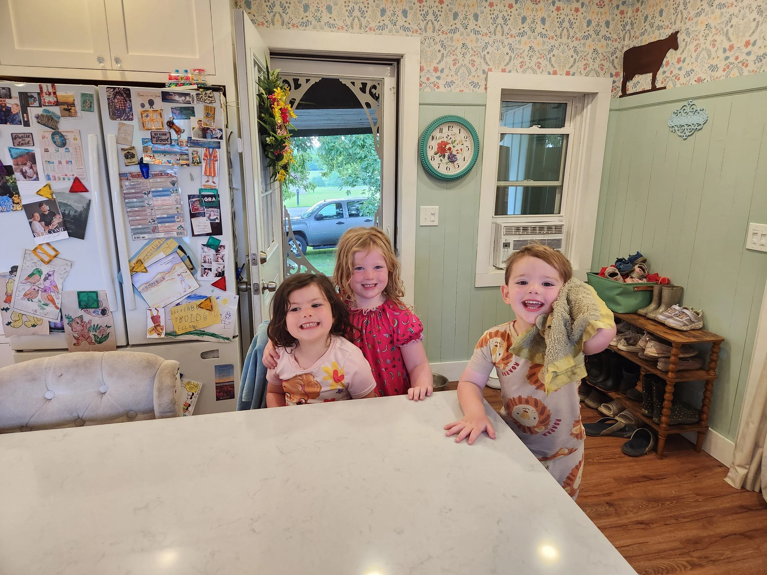 Four young children smiling at the camera while standing behind a kitchen counter in a cozy home. There are three girls and one boy, dressed casually, with a background showing a fridge covered with magnets and pictures, a window, and shoes on a rack.