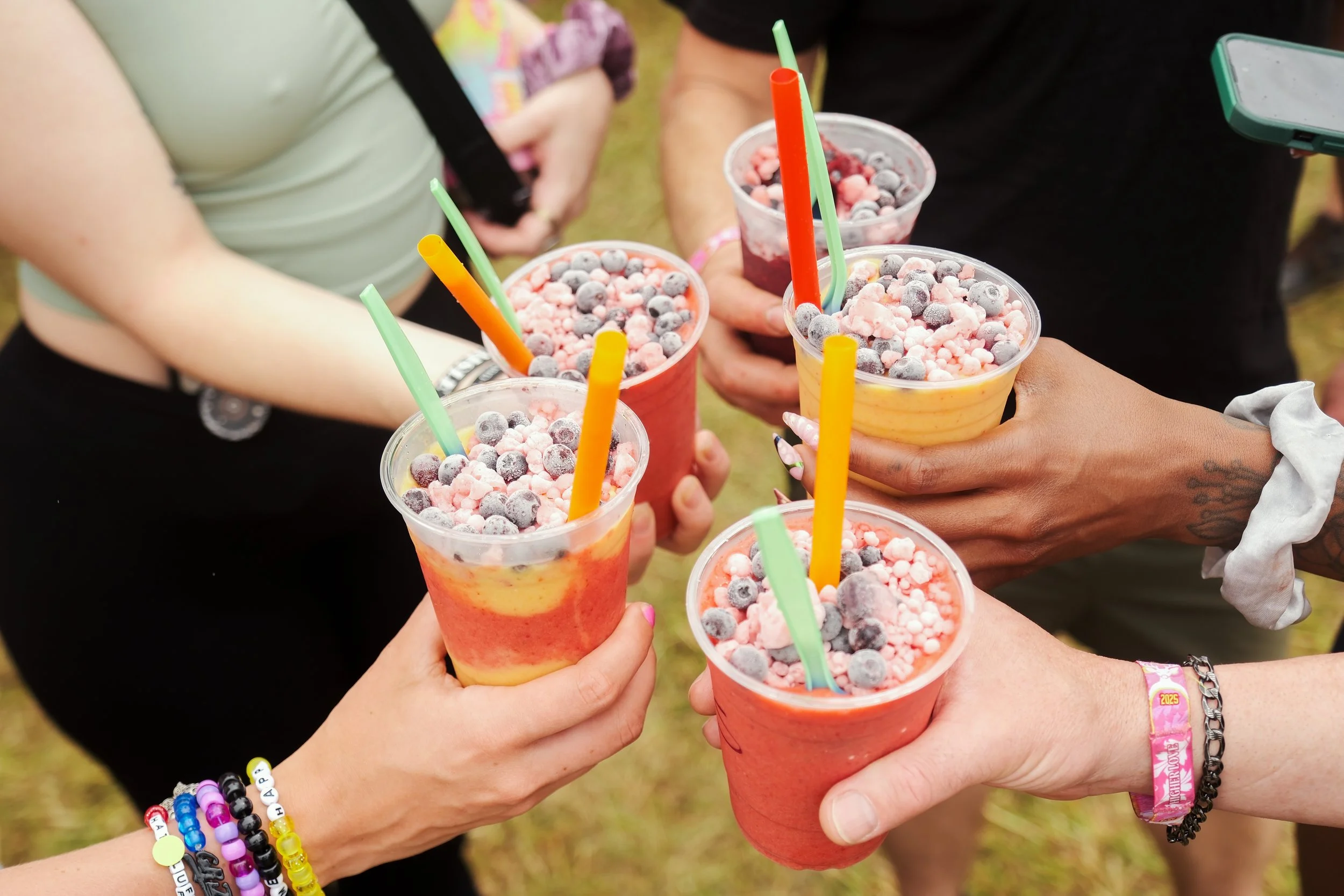 People holding colorful fruit smoothies topped with frozen berries and pink cereal outdoors