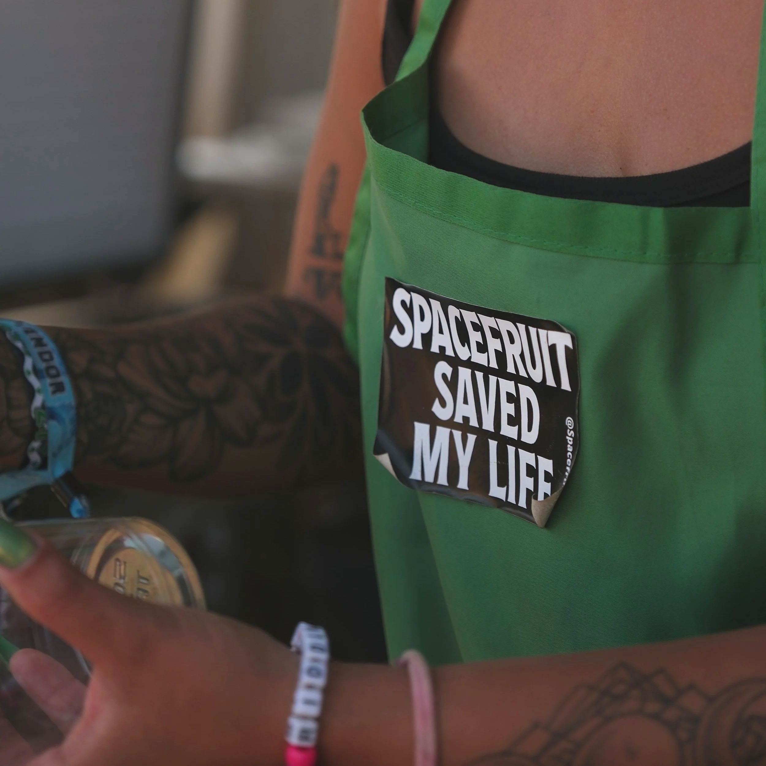 Close-up of a person wearing a green apron with a “Spacefruit Saved My Life” patch and holding a smartphone.