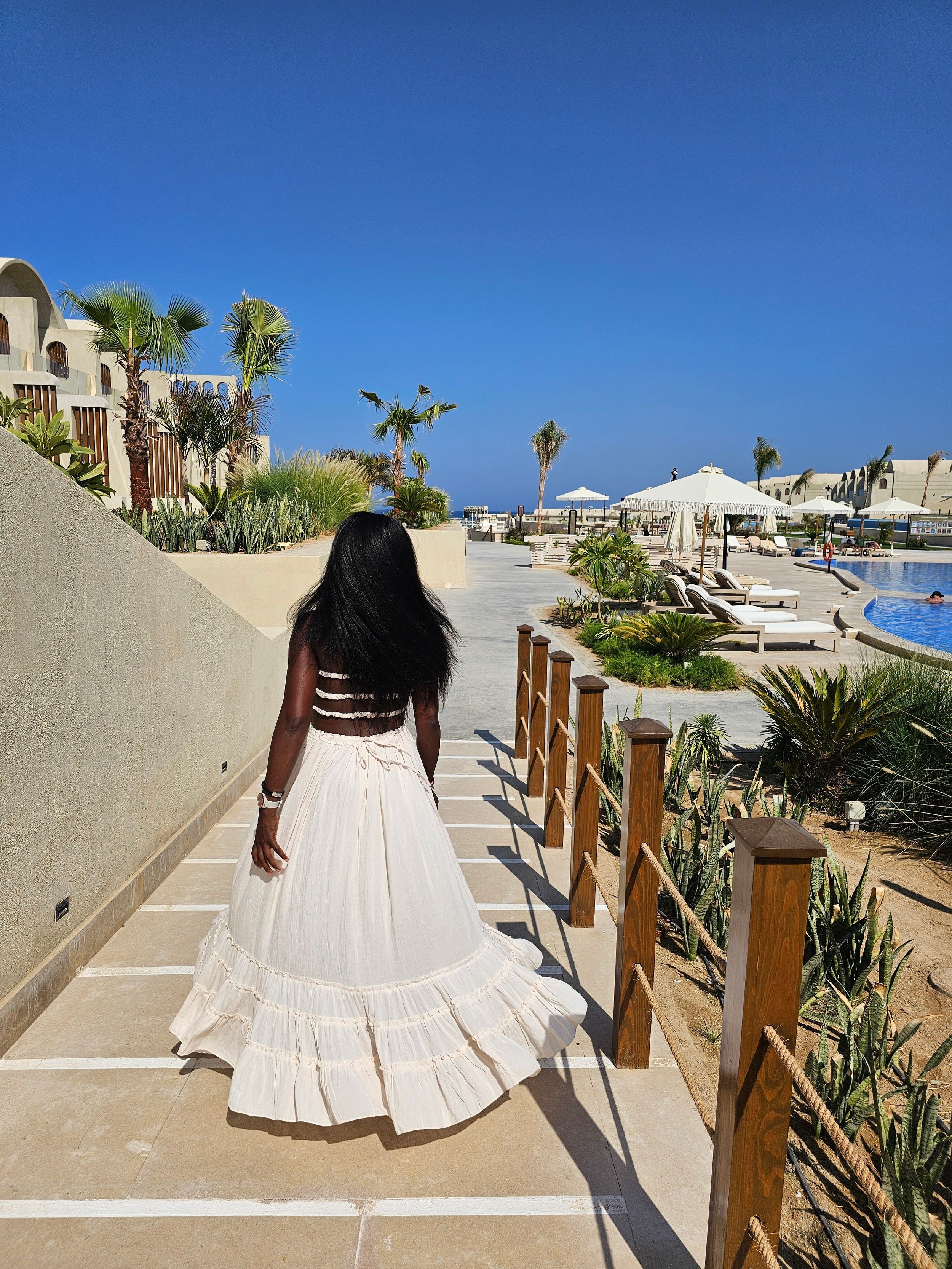 A woman with long dark hair wearing a flowing white dress walking along a pathway near a swimming pool area with lounge chairs, umbrellas, and palm trees under a clear blue sky.